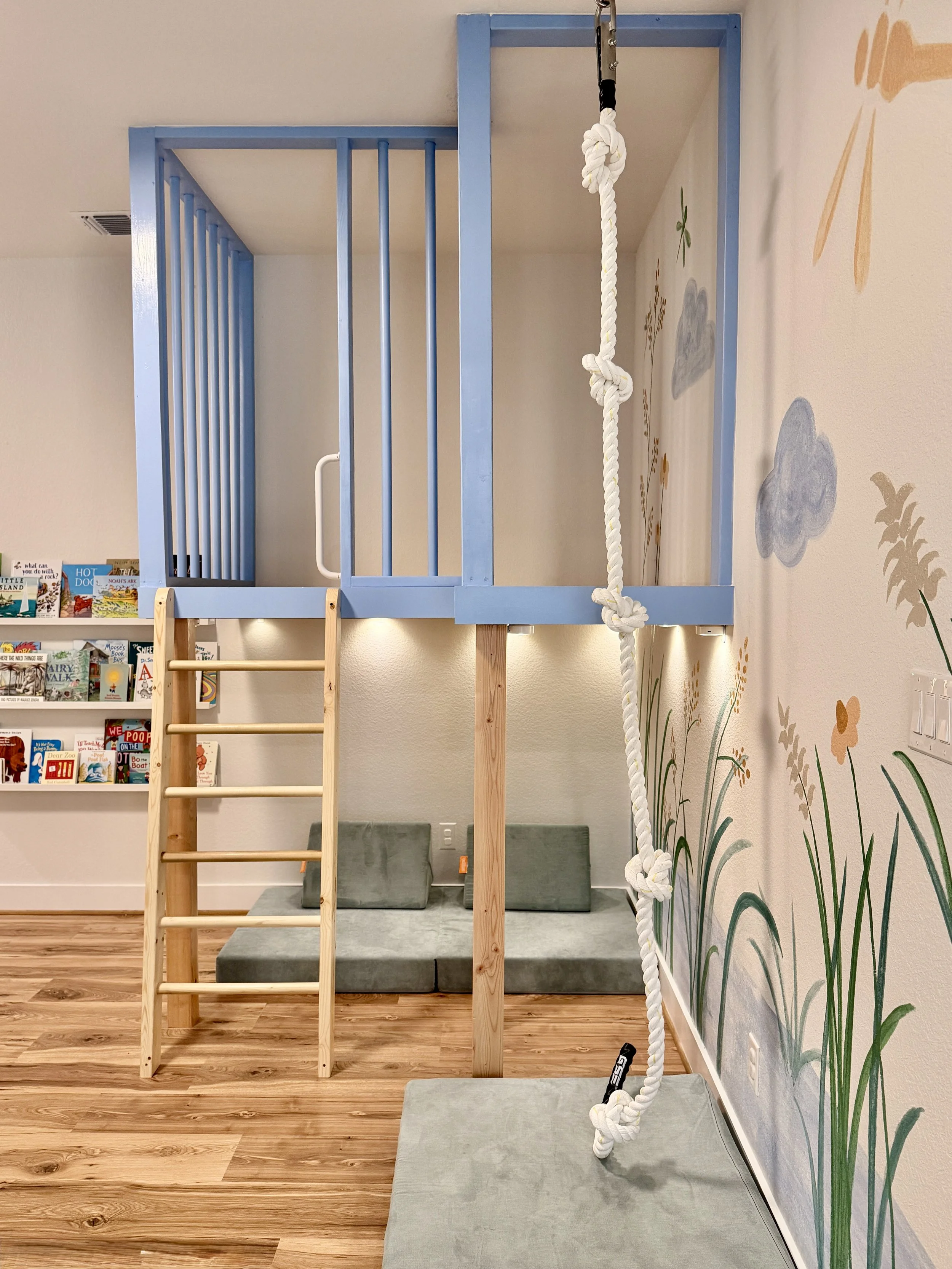 Blue play platform with a climbing rope exit, reading nook underneath and a whimsical mural on the wall