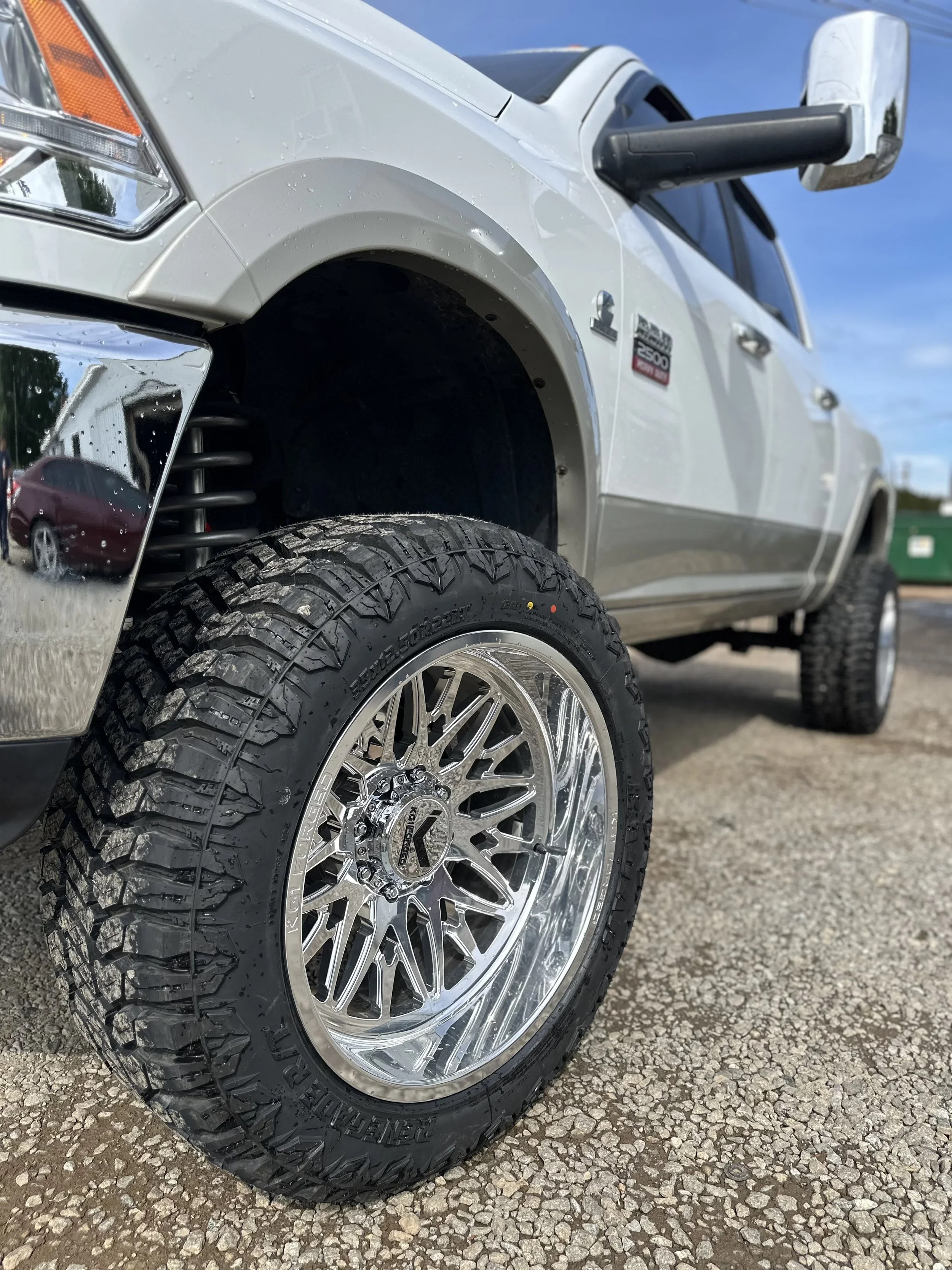 A stack of four large off-road tires next to black alloy wheels on a rusty floor against a black brick wall.