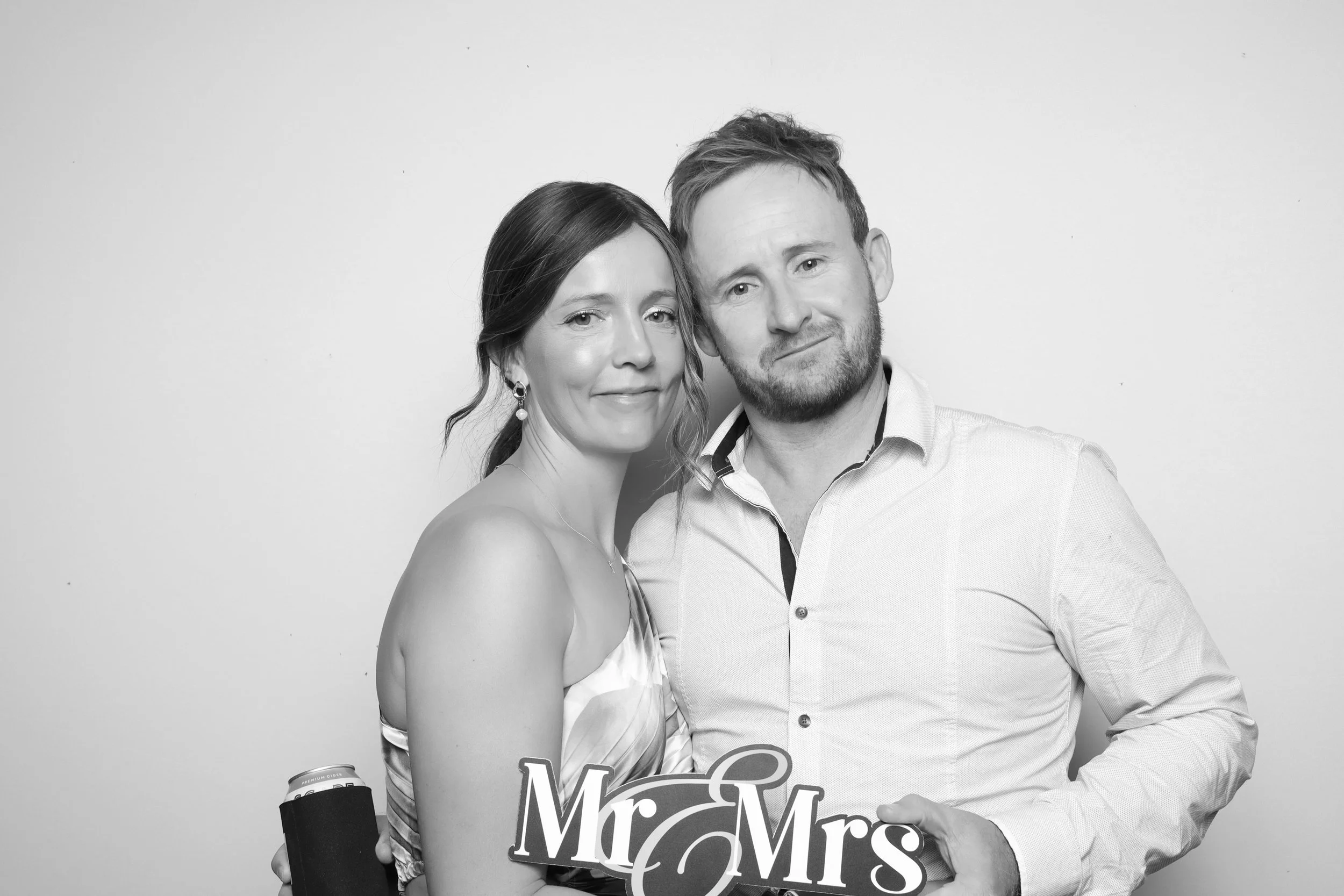 A black-and-white photo of a smiling woman and man standing close together against a plain white background. The woman is holding a sign that reads "Mr & Mrs" and has her head slightly tilted towards the man. The man is wearing a light-colored button-up shirt and has a beard. The woman has earrings and is dressed in a sleeveless top.