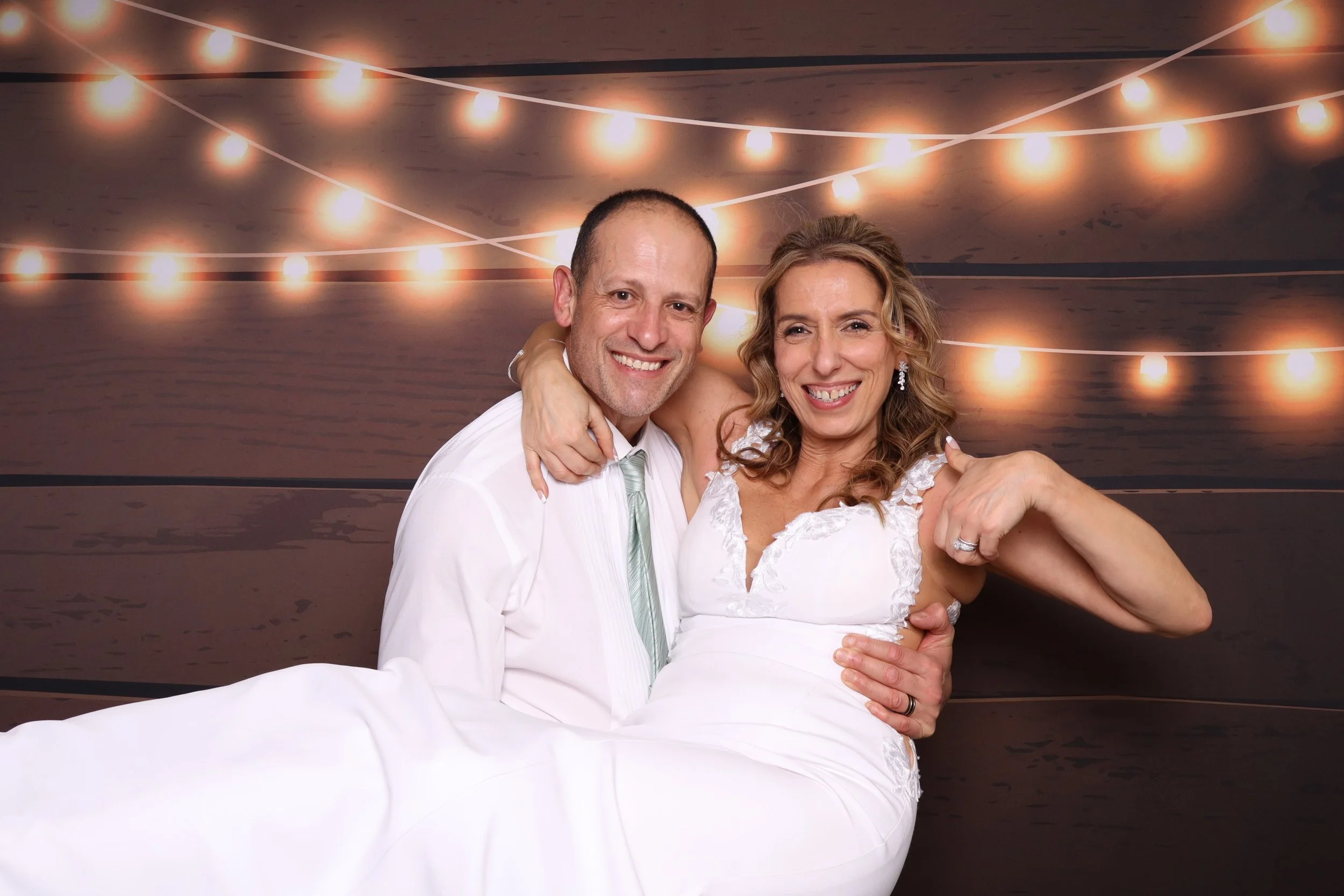 A happy couple, dressed in wedding attire, smiling and embracing each other in front of a background with string lights reflected on water.