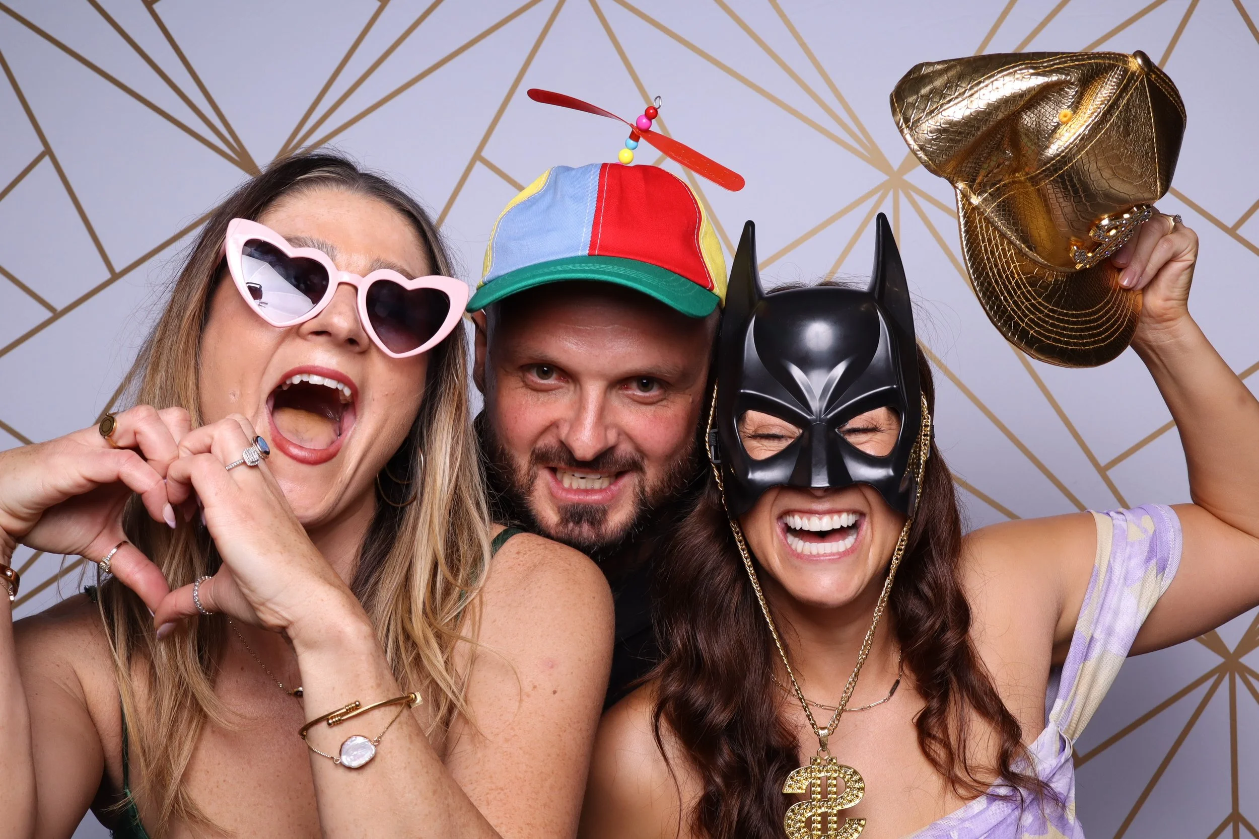 Three people celebrating with fun accessories: woman with pink heart-shaped sunglasses, man with a colorful baseball cap and a toy dragonfly hovering above, woman with a black mask and gold purse, against a geometric patterned backdrop.