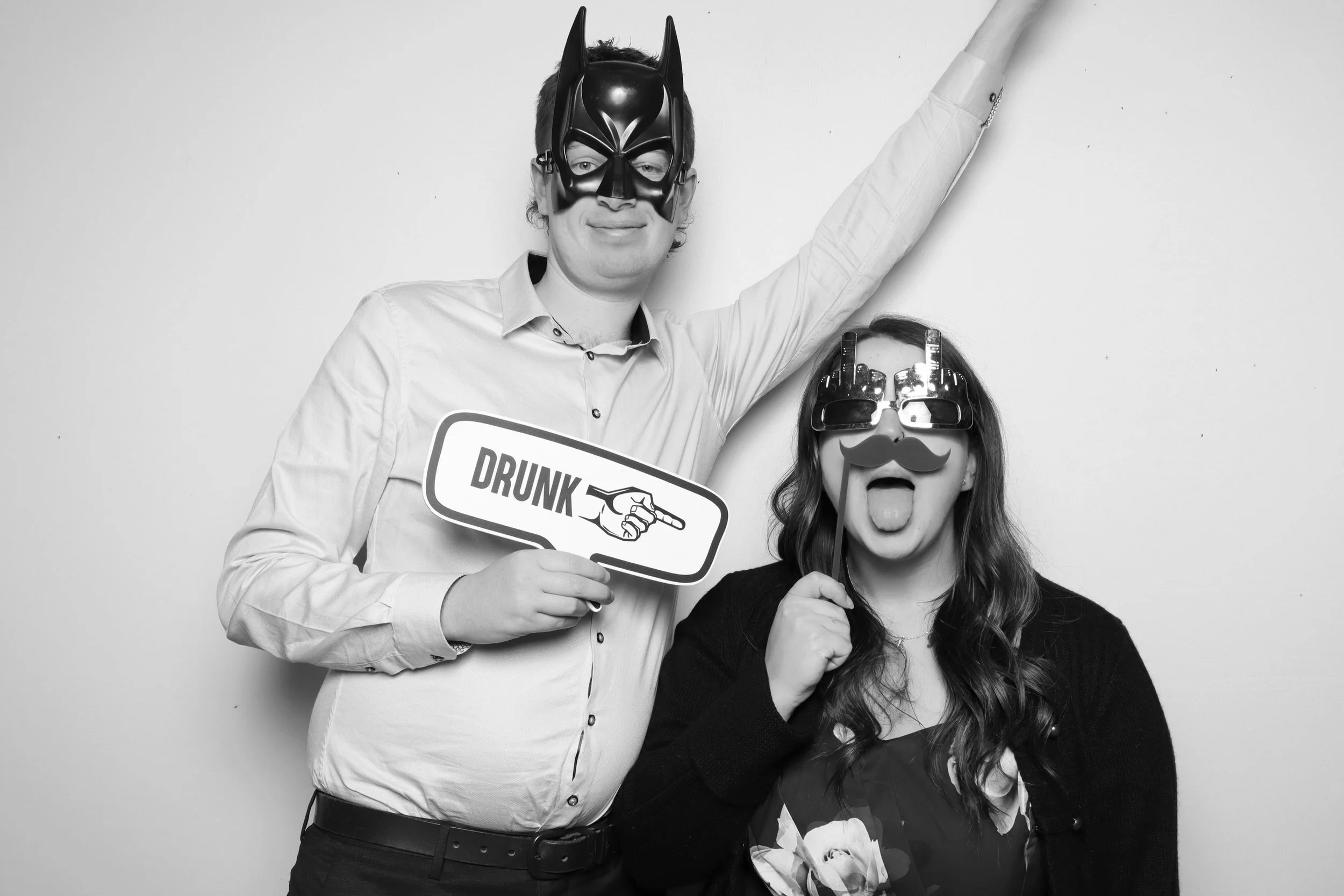 A man and woman are dressed in humorous costumes and masks. The man is wearing a Batman mask and holding a sign that says 'DRUNK' with an arrow pointing to the right, and the woman is wearing oversized sunglasses with a fake mustache and sticking her tongue out.