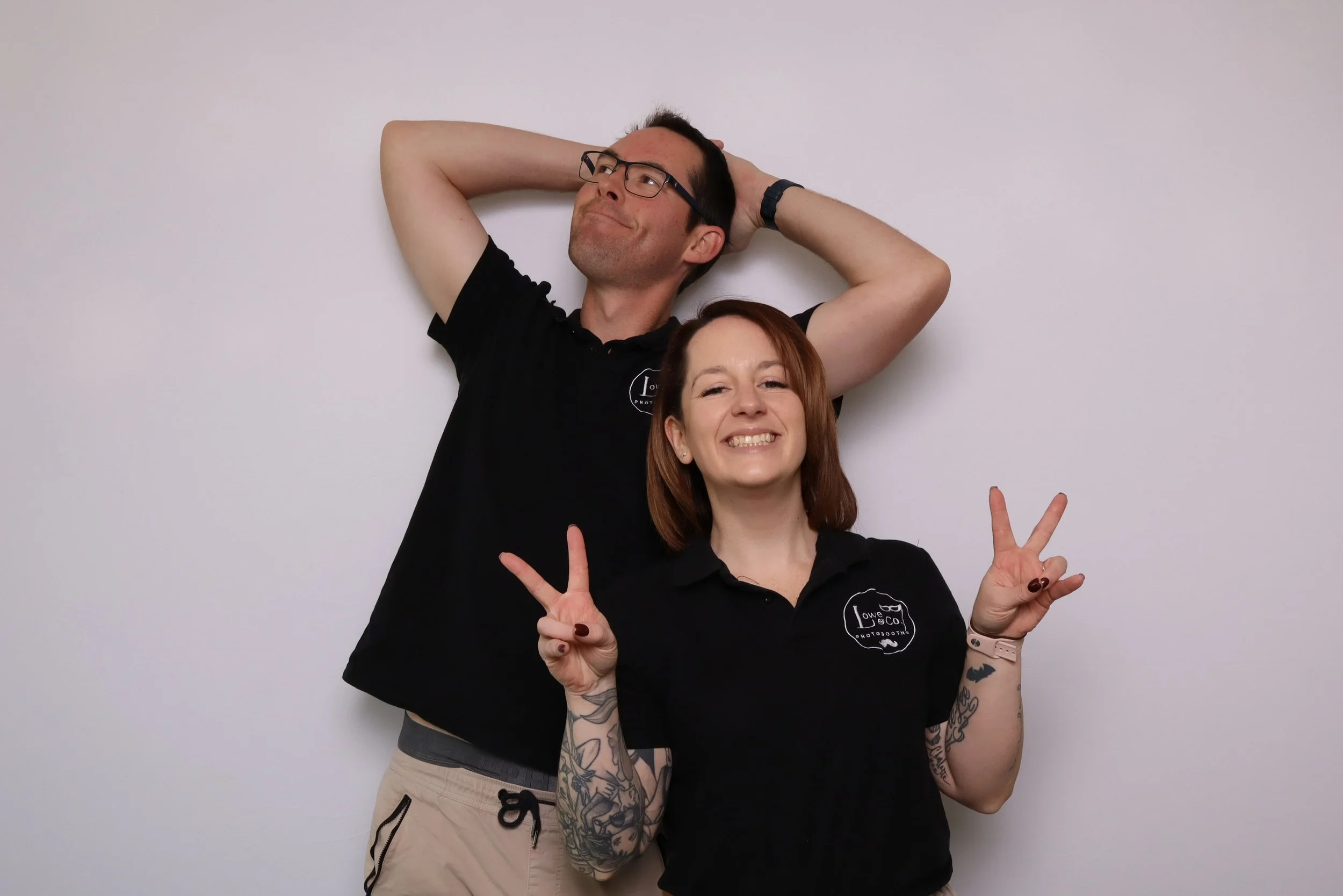 Two smiling people posing in front of a plain white wall, inside the photobooth, making peace signs with their hands, and standing closely together.