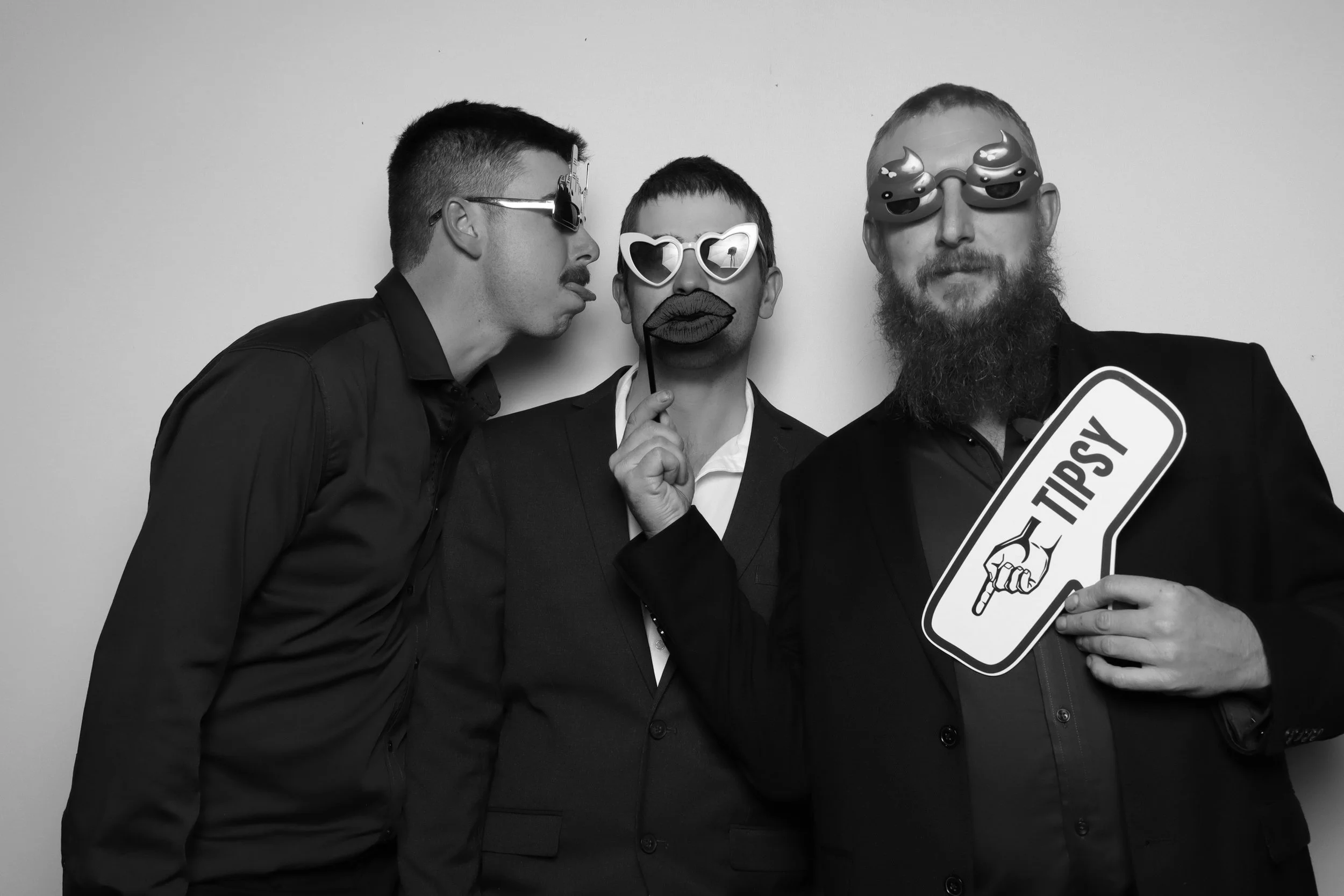 Group of three people wearing fun sunglasses and holding props, including a stick with lips, a sign that says 'TIPSY,' and glasses with cartoonish eyes, in front of a plain wall.