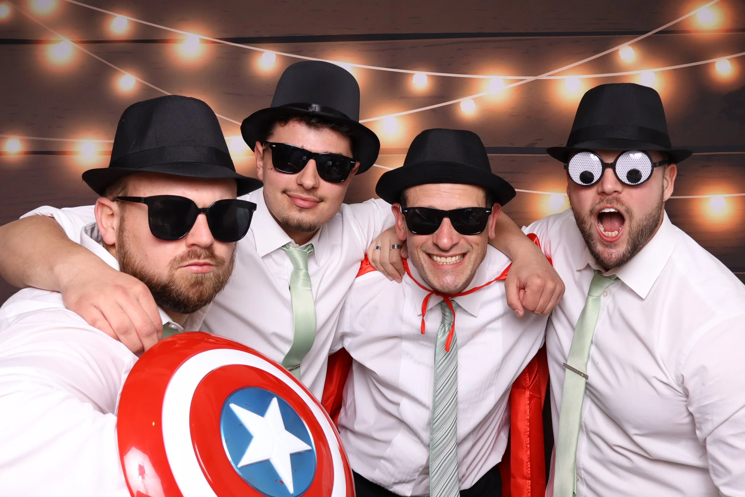 Four men wearing black hats, sunglasses, and white shirts with ties, posing together at a celebration with string lights overhead, one holding a Captain America shield and balloon.