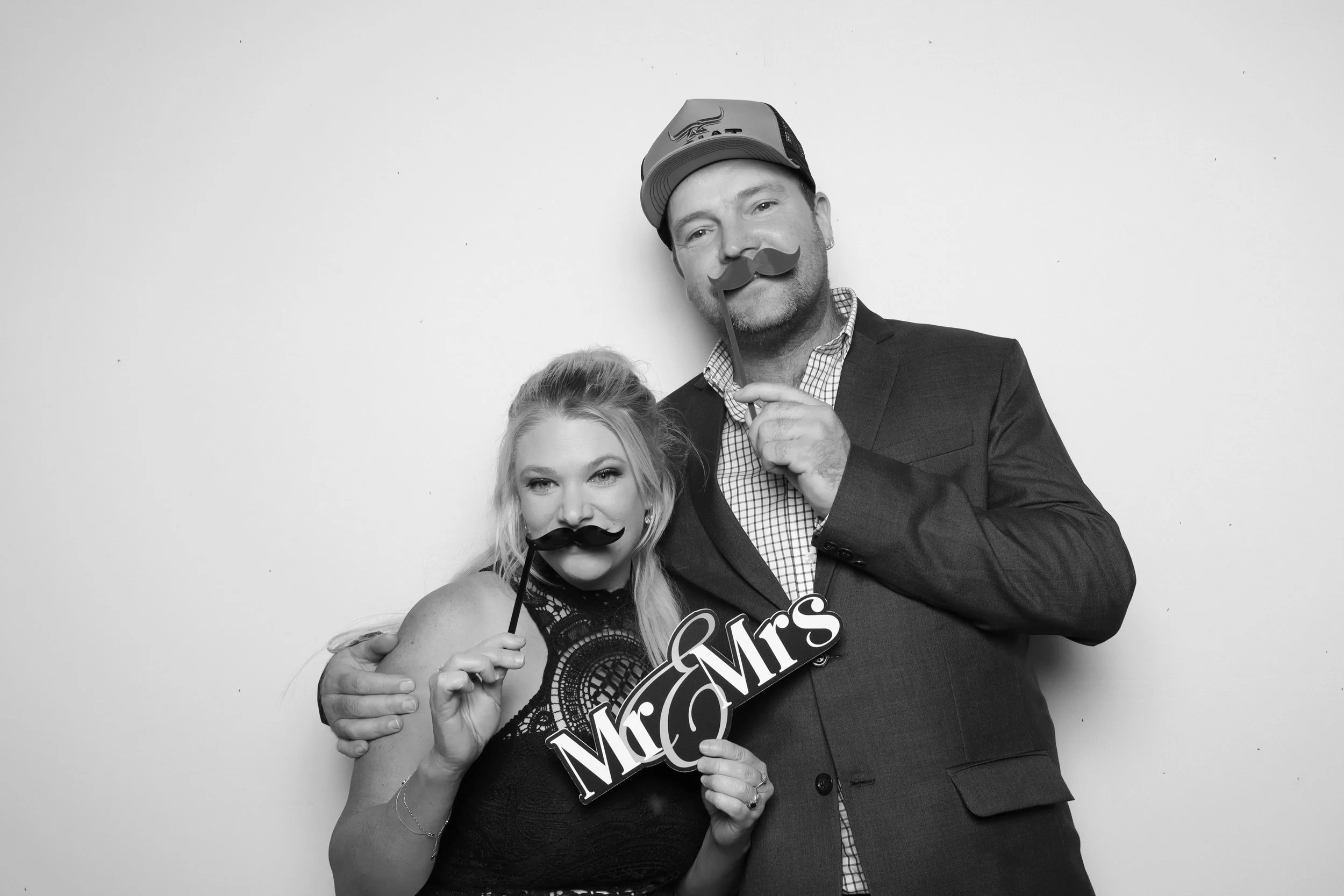 A man and woman at a celebratory event, both holding mustache props. The woman is wearing a lace dress, and the man is dressed in a suit and checkered shirt, with a cap. The man is also holding a sign that says 'Mrs.' The photo is in black and white.