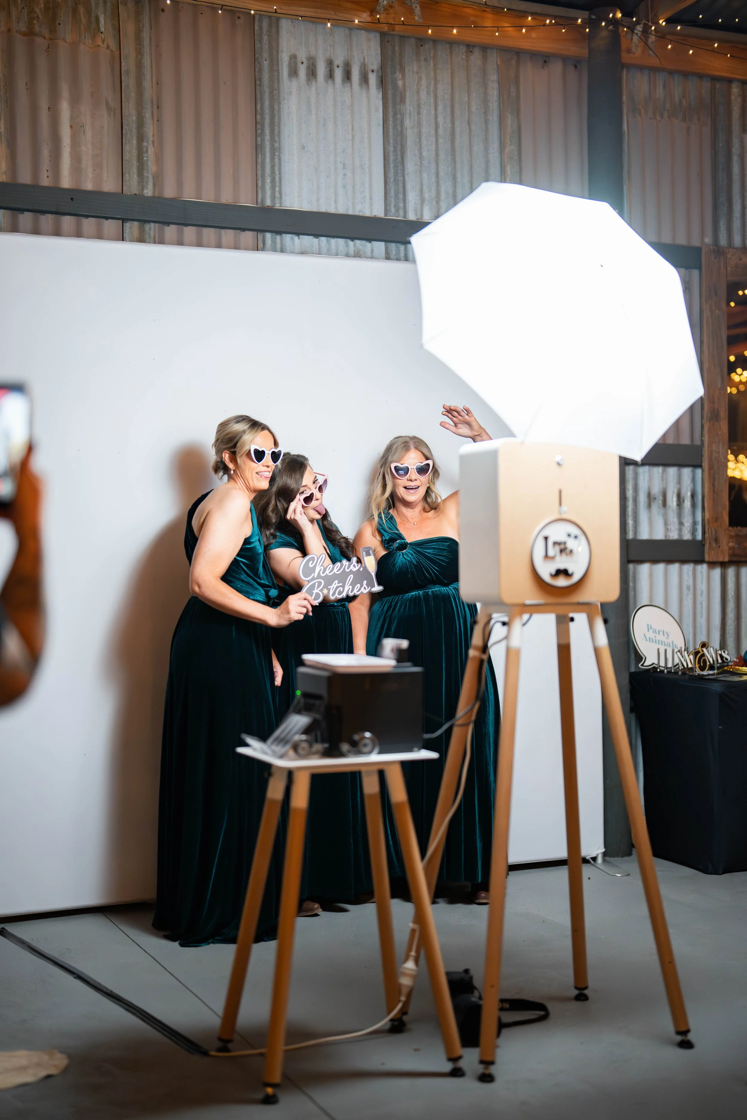 Four women in evening dresses and large heart-shaped sunglasses participate in a photo booth session at a party, with some holding props. They are behind a white photo backdrop and there is professional lighting equipment set up.