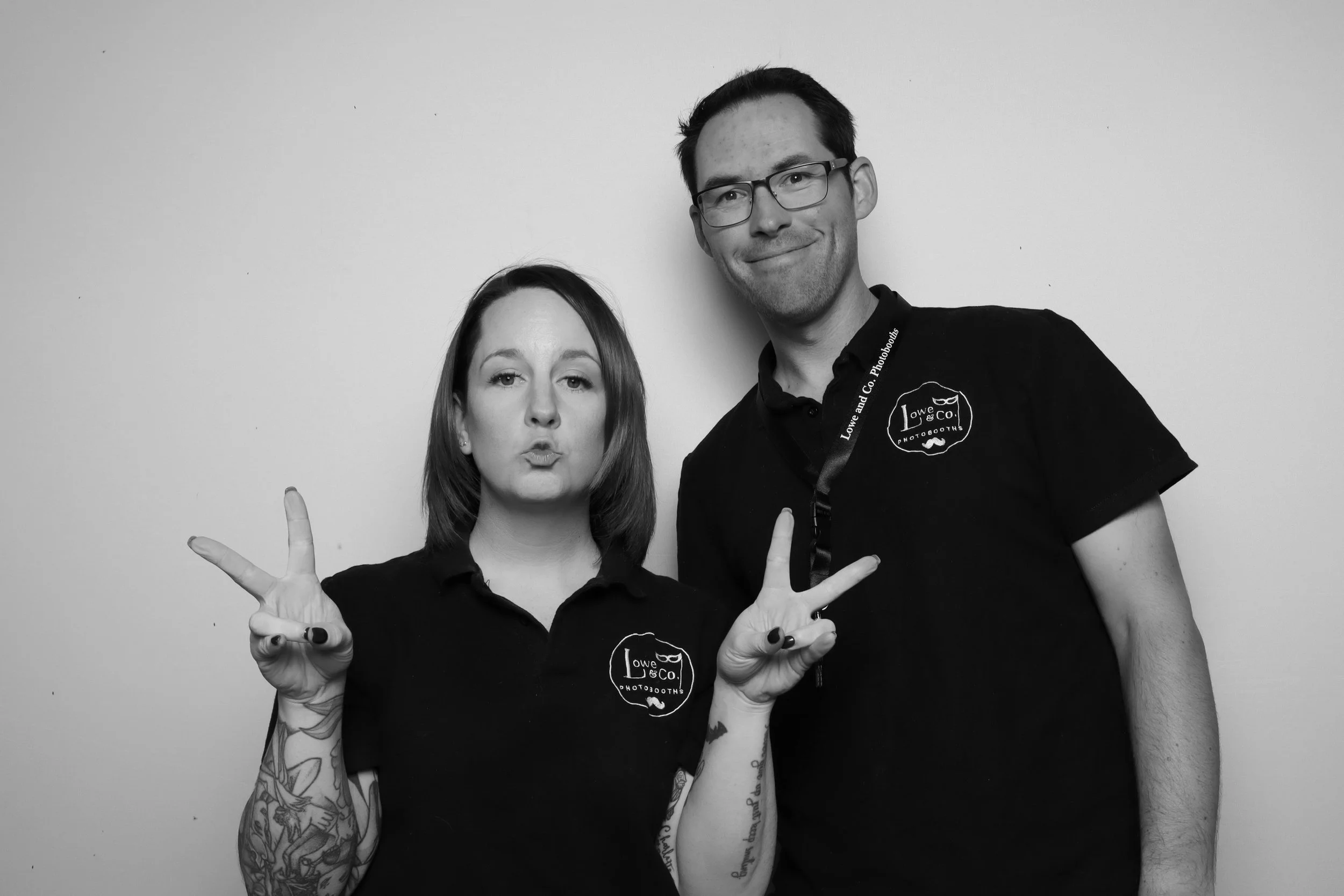 A black-and-white photo of a woman and a man posing together against a plain wall. The woman has shoulder-length hair, making a peace sign with both hands, and is wearing a dark shirt. The man has short dark hair, glasses, and is smiling, wearing a black polo shirt with a stitched logo on the chest that says "Lowe and Co. Photobooths." They seem to be at a fun event or photo booth.