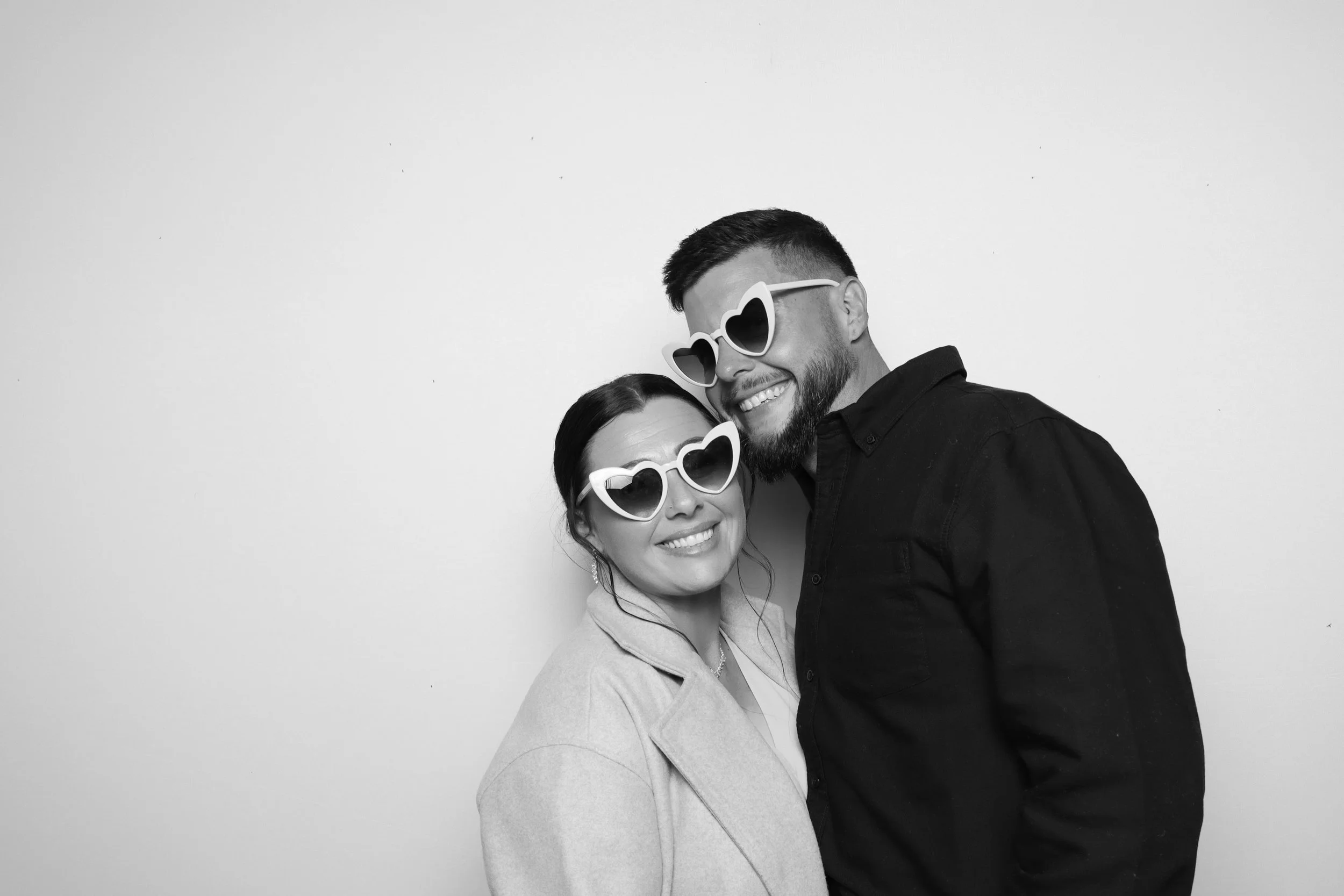 A smiling woman and man wearing heart-shaped sunglasses, hugging each other against a plain white wall.