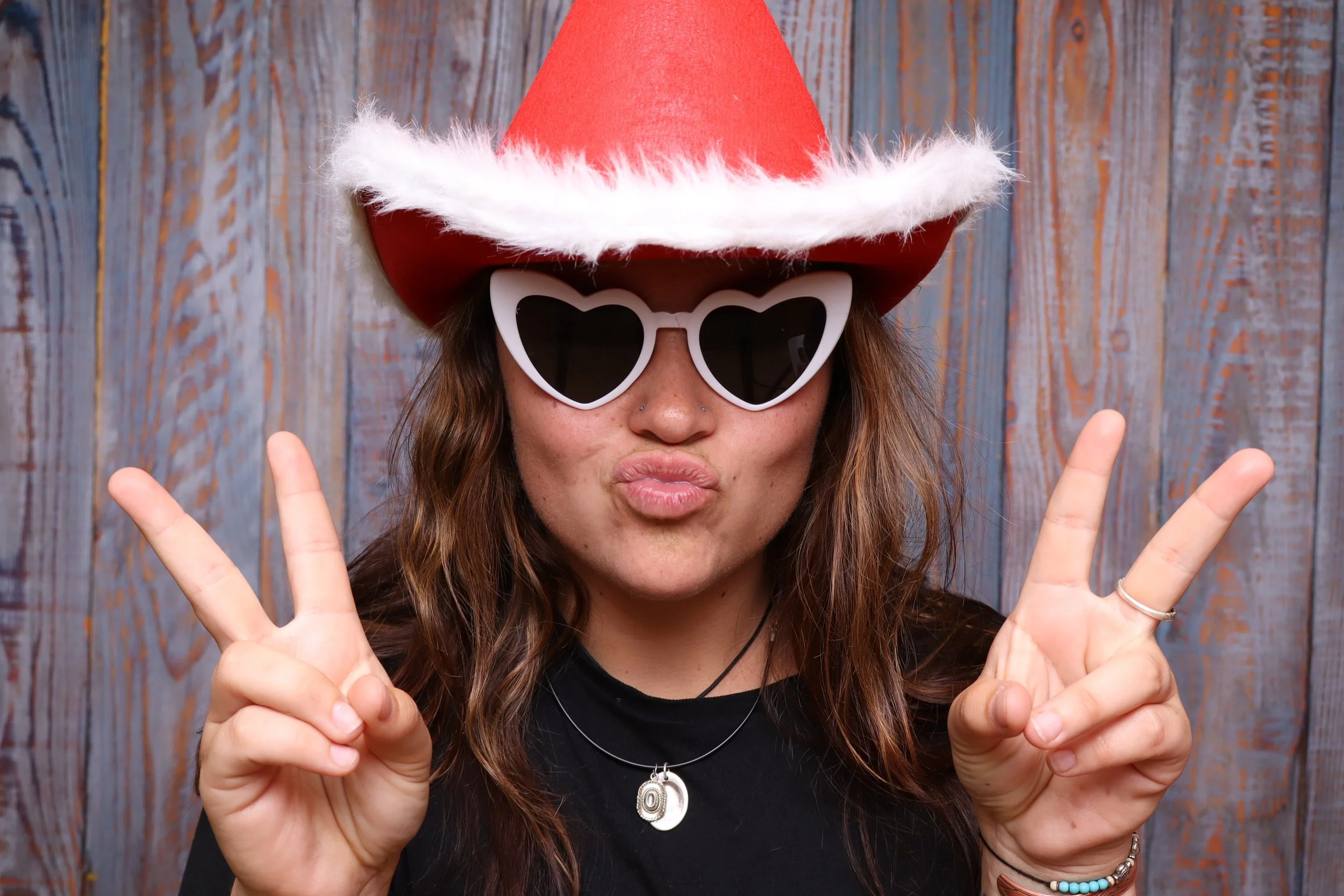 Woman inside a photobooth wearing a red Santa hat with white trim, white heart-shaped sunglasses, and making a peace sign with her fingers in front of a wooden background.