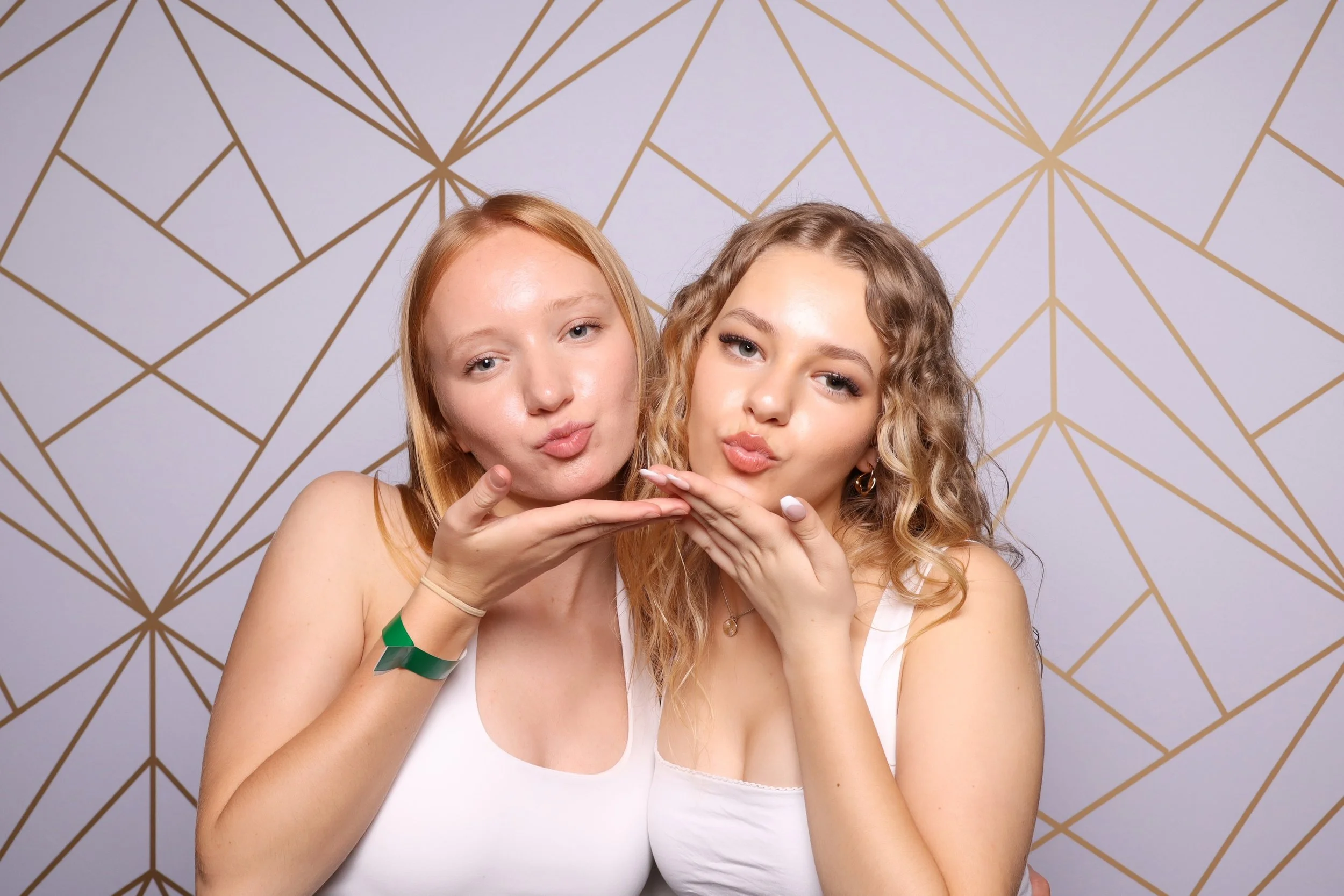 Two young women with blonde and curly hair posing with hands under their chins in front of a gold geometric patterned wall inside the photobooth in Melbourne.