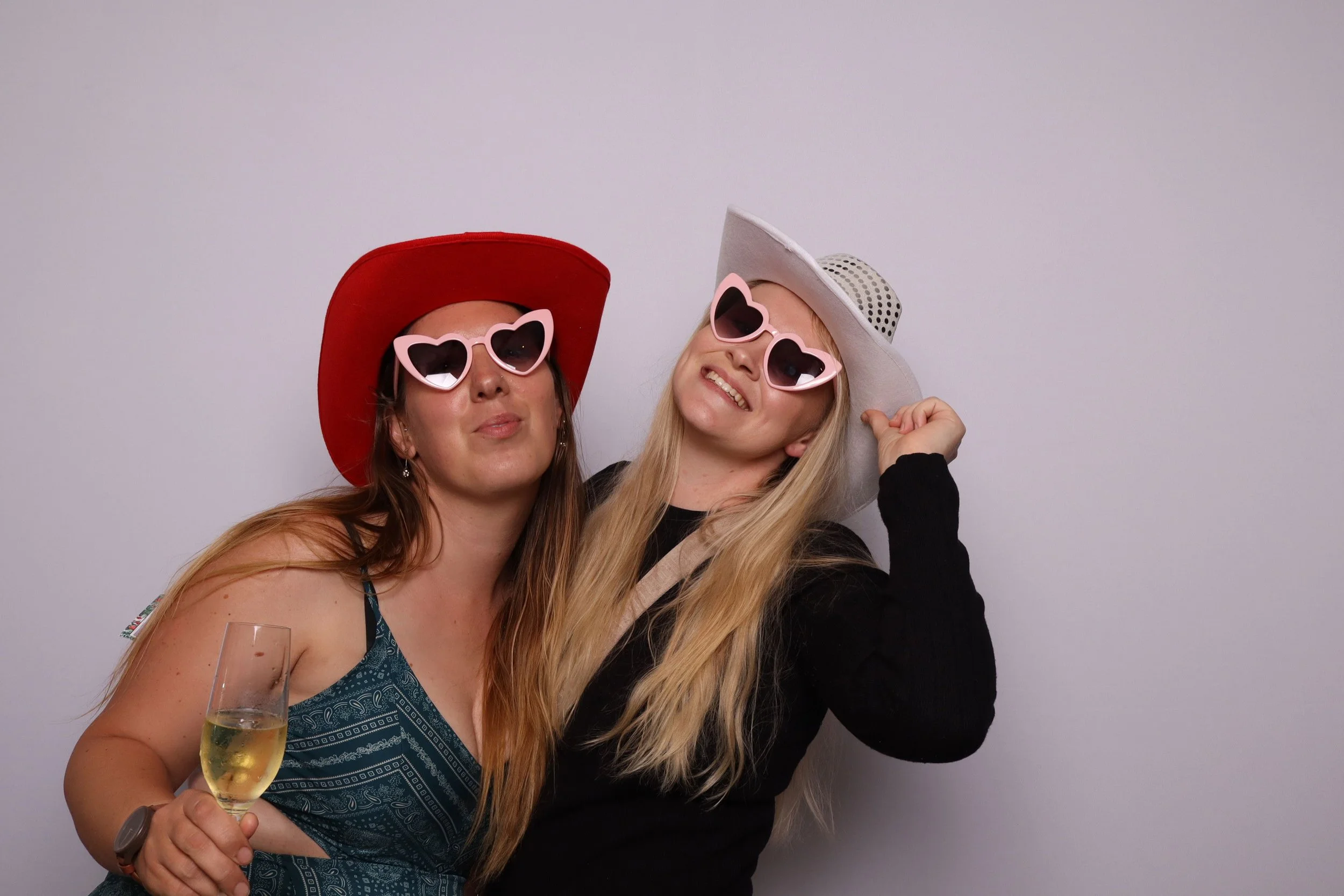 Two women inside the booth, wearing large hats and pink heart sunglasses, smiling and posing together. One woman is holding a glass of champagne.