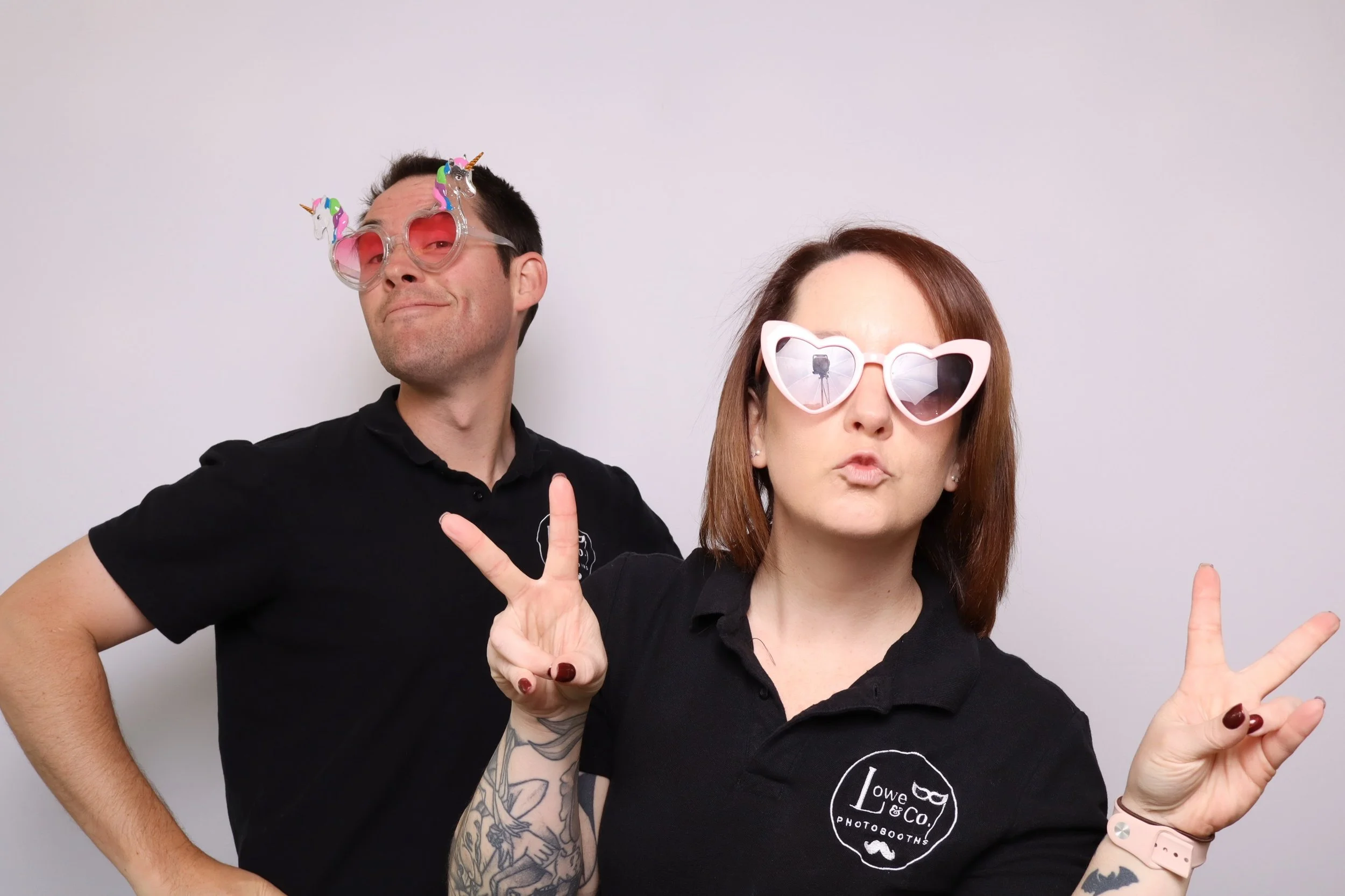 Two people posing in front of a plain white background, wearing fun sunglasses with unicorn and heart shapes, making peace signs and playful expressions.