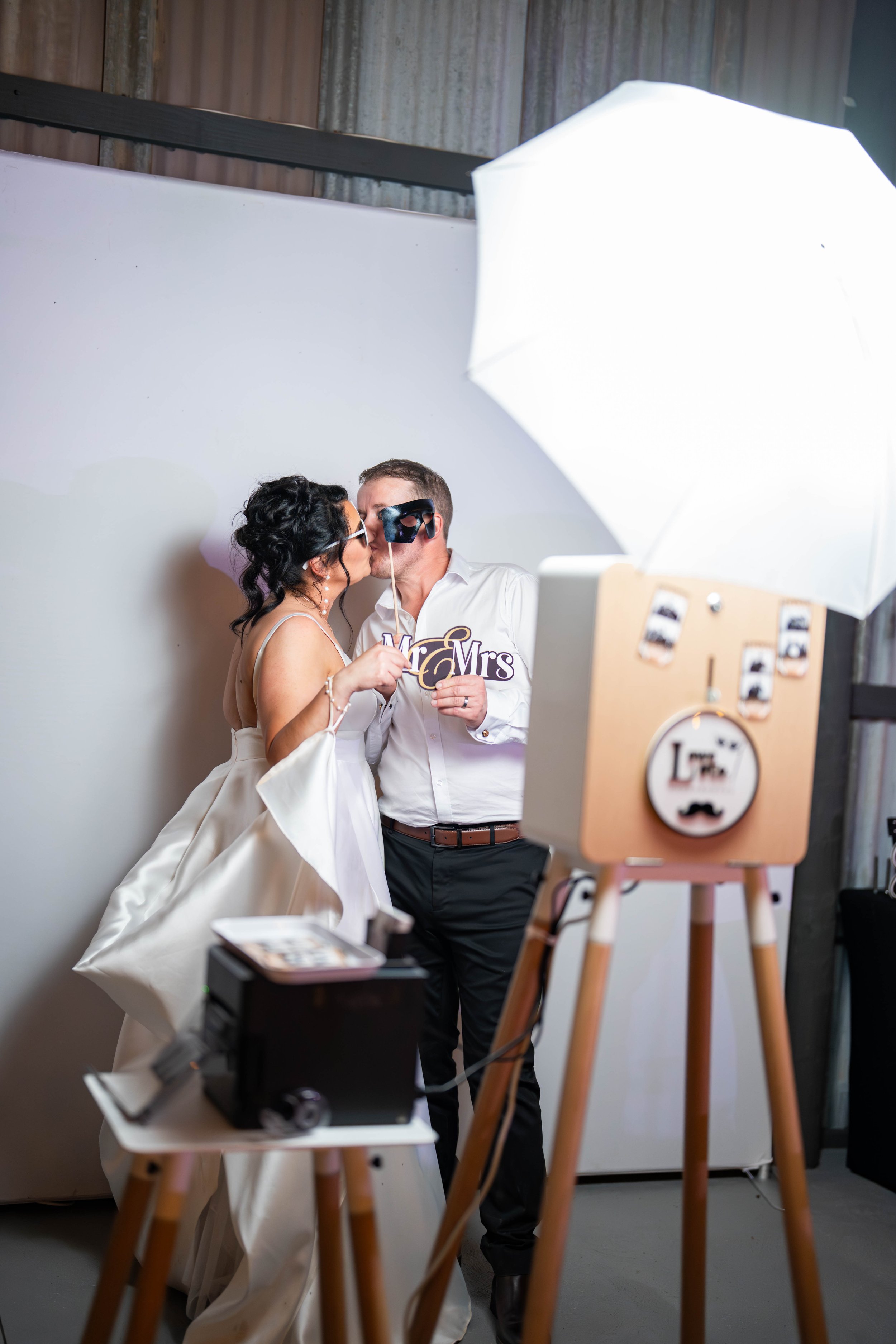 A man and woman in wedding attire sharing a kiss in a photo booth at a wedding reception, with a camera, photo props, and a white umbrella in the background.