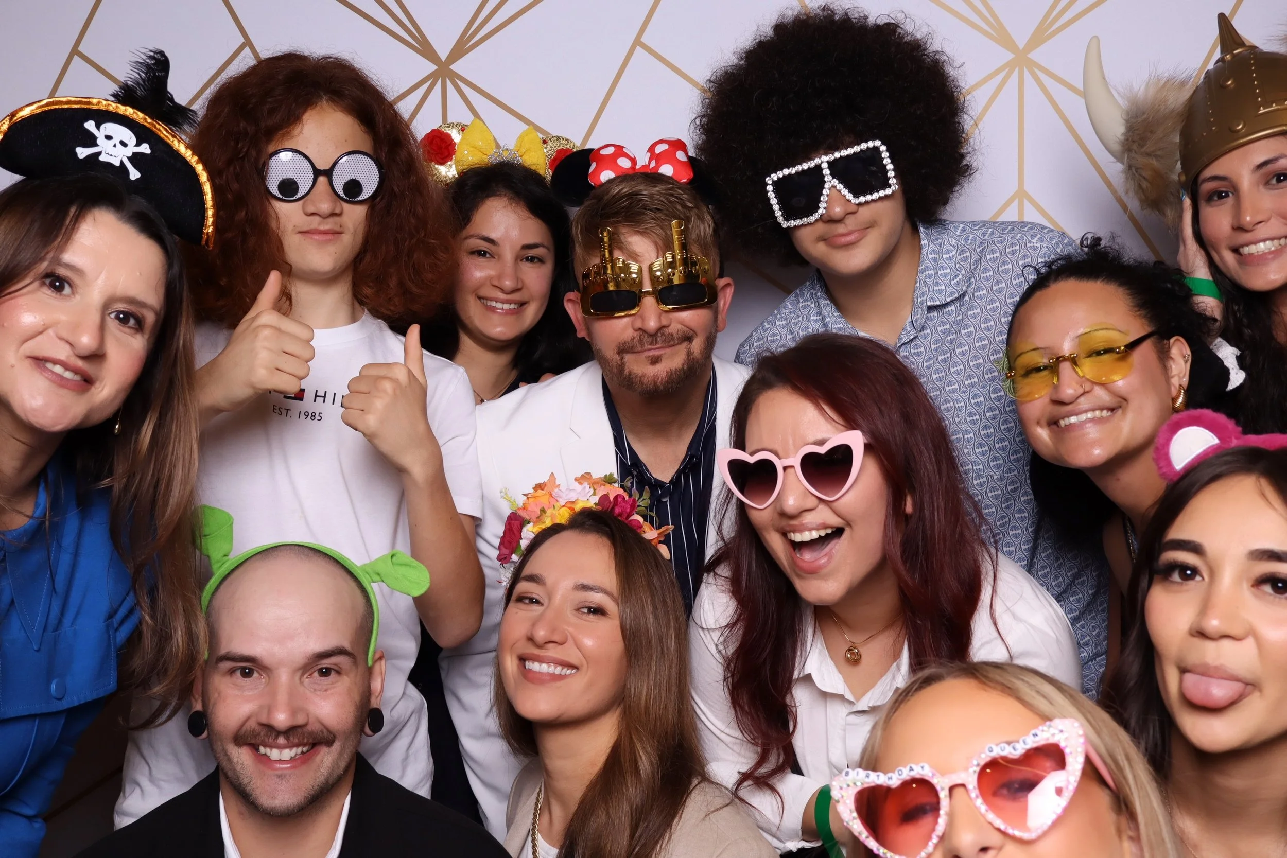 Multiple people wearing fun and colorful costumes and accessories, smiling and posing for a group photo in front of a geometric-patterned background.
