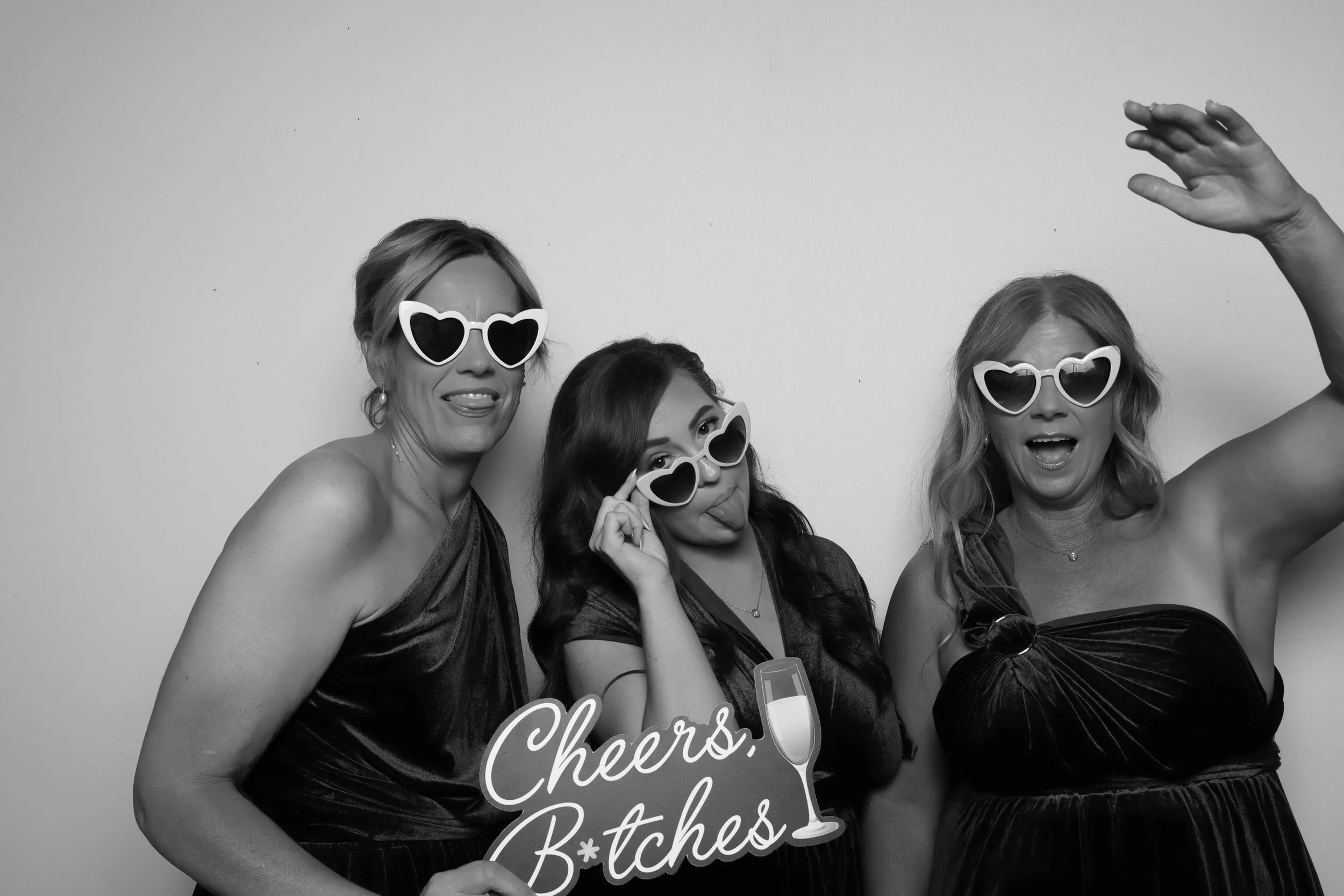 Black and white photo of three women at a party, wearing heart-shaped sunglasses, dressed in elegant dresses, holding a sign that says "Cheers!" and "Bitches." They appear to be celebrating and having fun, with one woman raising her hand in the air.