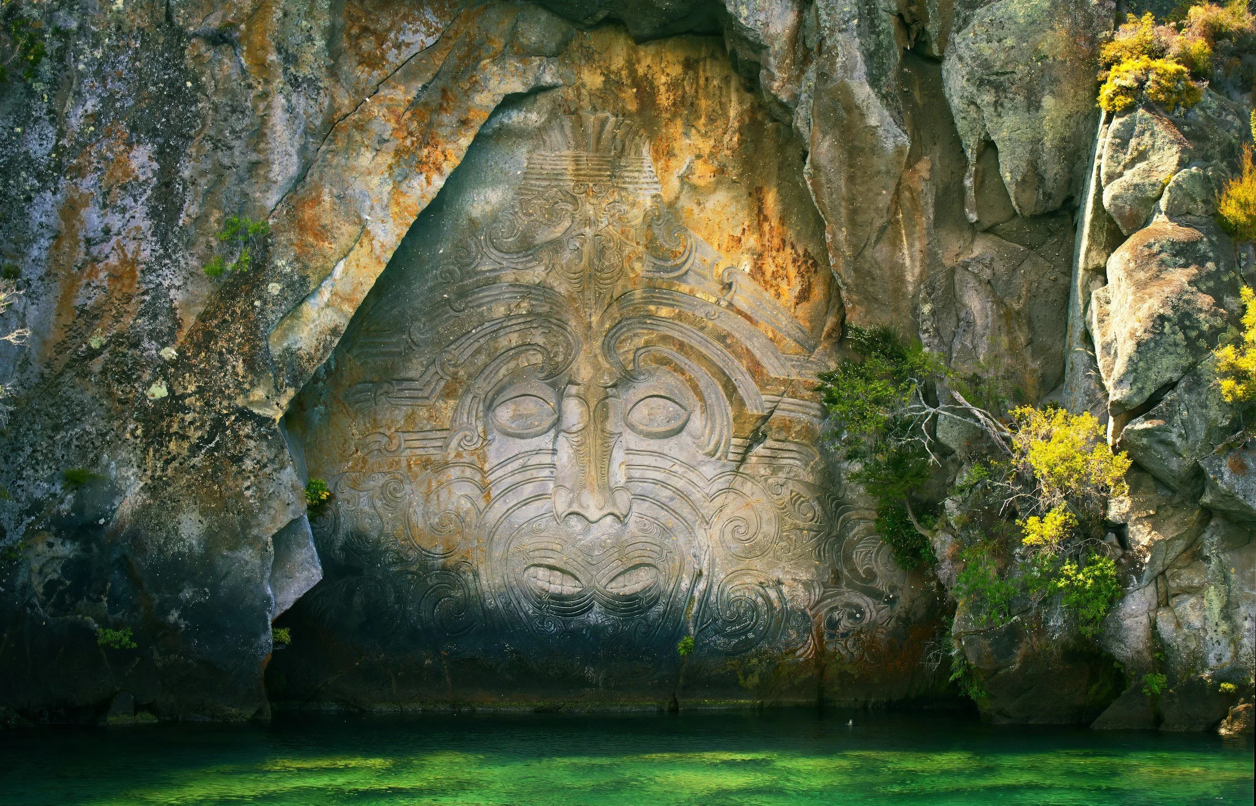 A carved stone face on a cliffside surrounded by yellow and green foliage, reflecting in a body of water below.