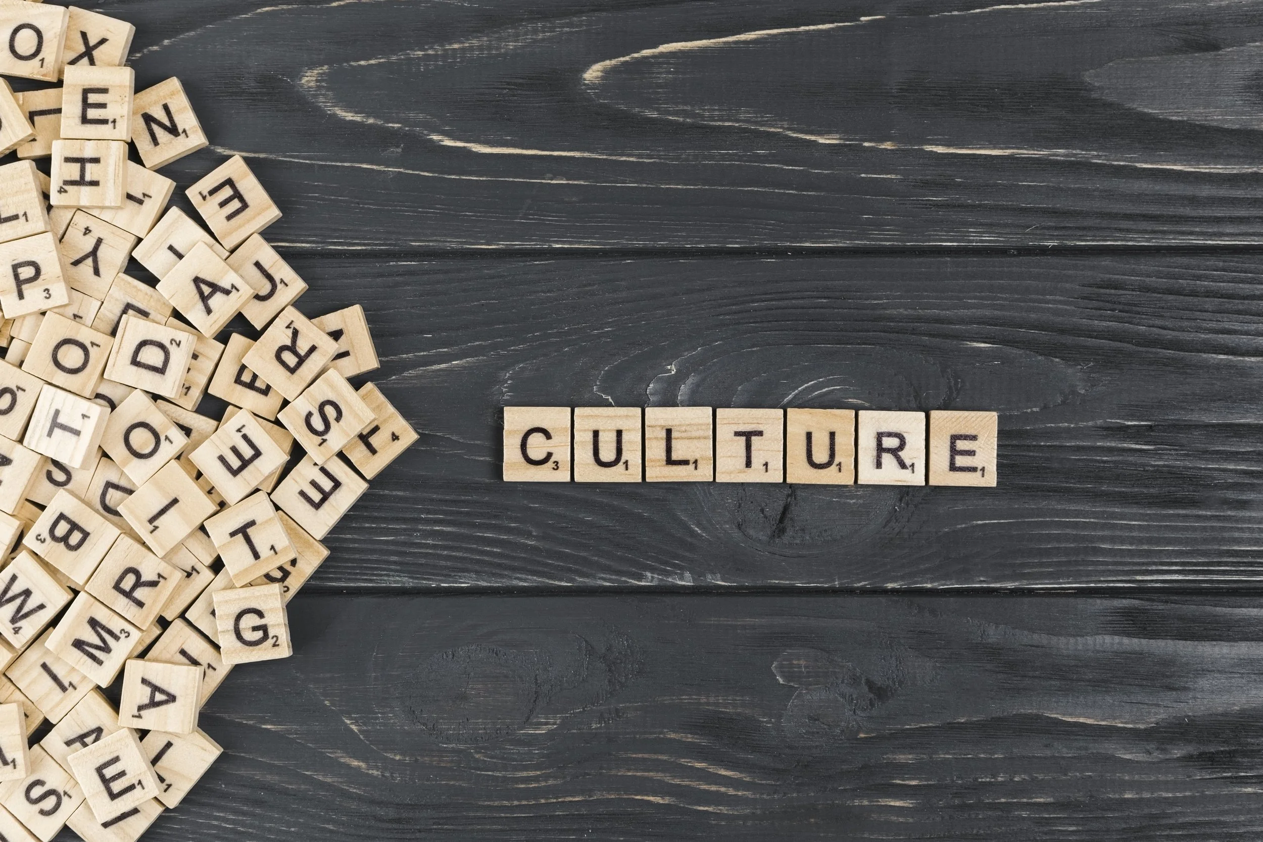 Scrabble tiles spelling out 'CULTURE' on a dark wooden surface, with a pile of scattered tiles to the left.