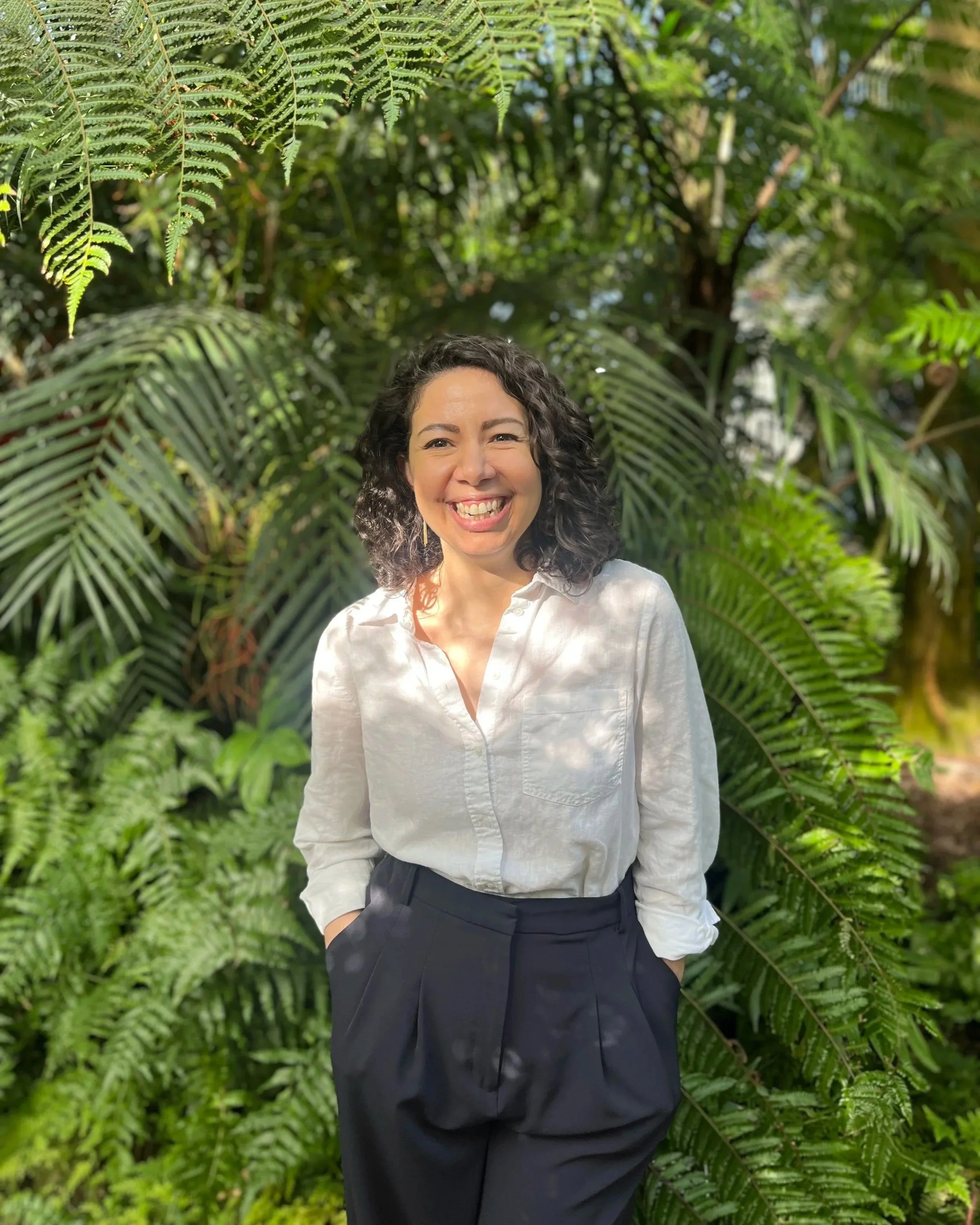 A woman with curly dark hair, smiling and wearing a white blouse and black pants, standing in front of lush green ferns and tropical plants.