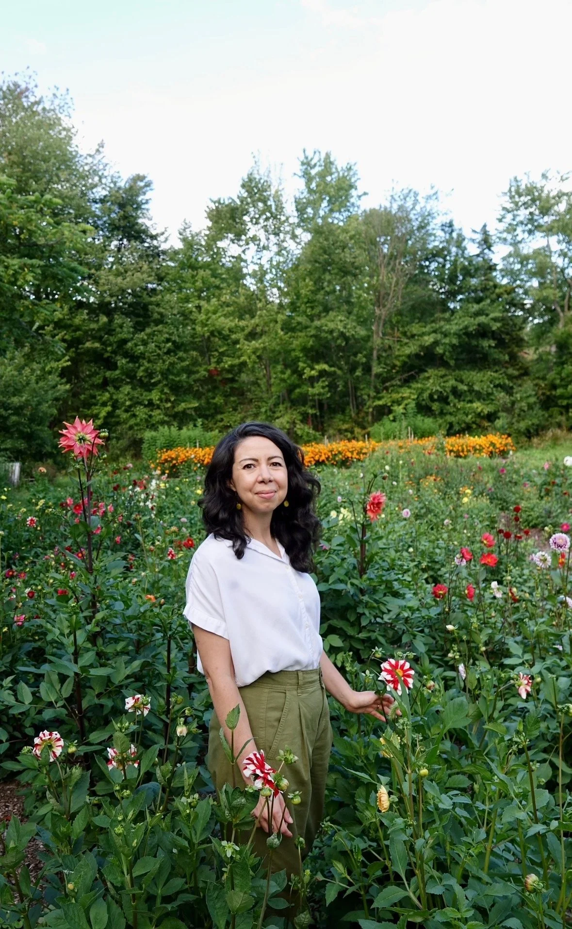 A woman with dark wavy hair standing in a colorful flower garden with tall green trees in the background.