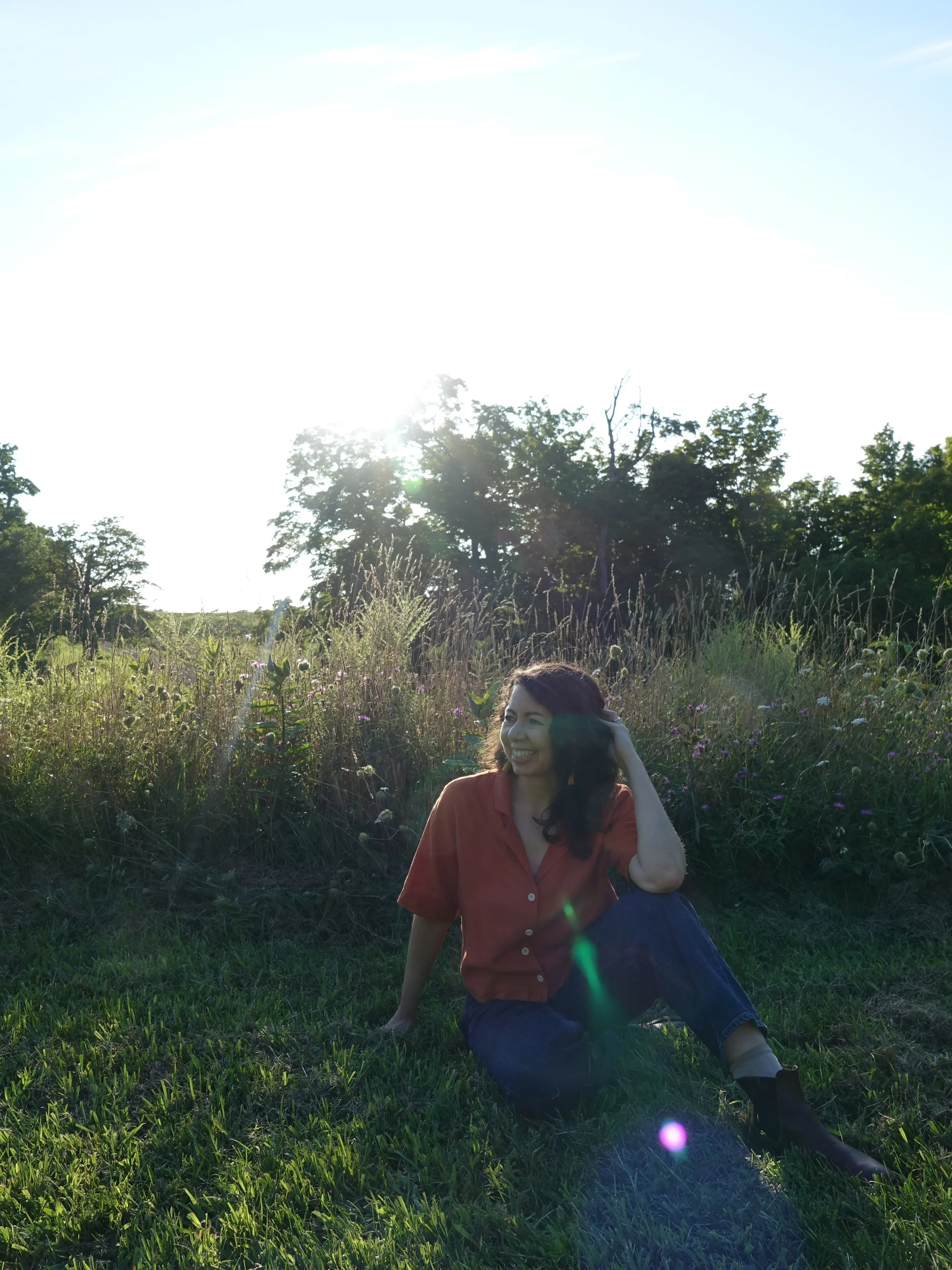 A woman sitting on grass in a field with wildflowers, smiling, wearing a red shirt and blue jeans, during a sunny day with trees in the background.
