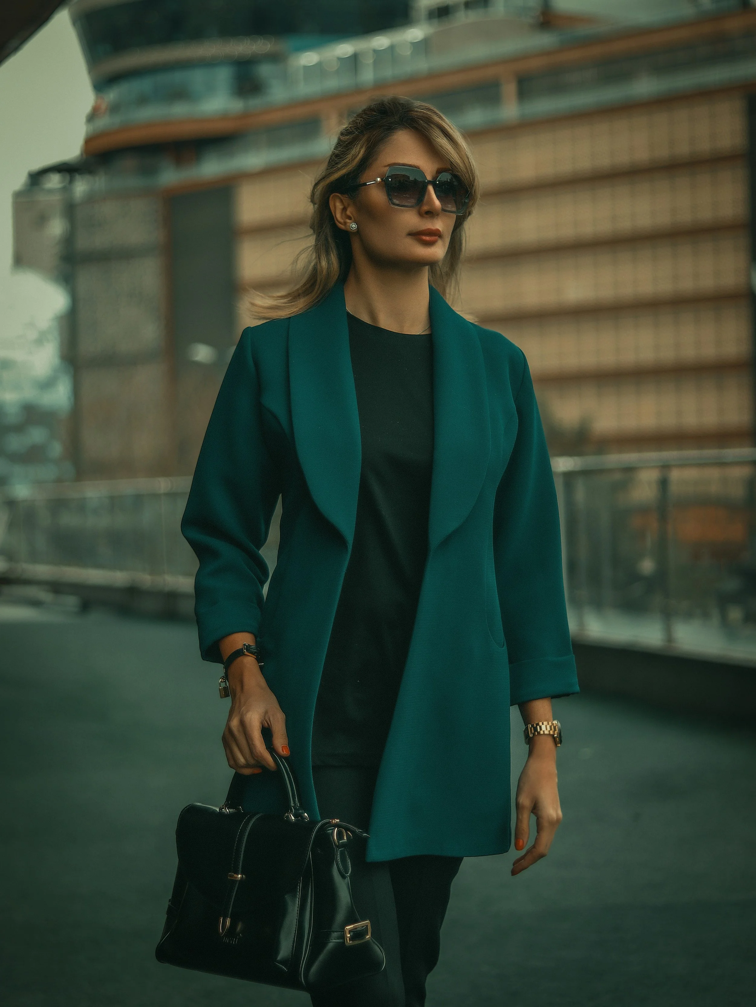 A woman with shoulder-length hair wearing large black sunglasses, a teal blazer, a black top, holding a black handbag, and wearing gold jewelry, standing outdoors in an urban setting with modern buildings in the background.