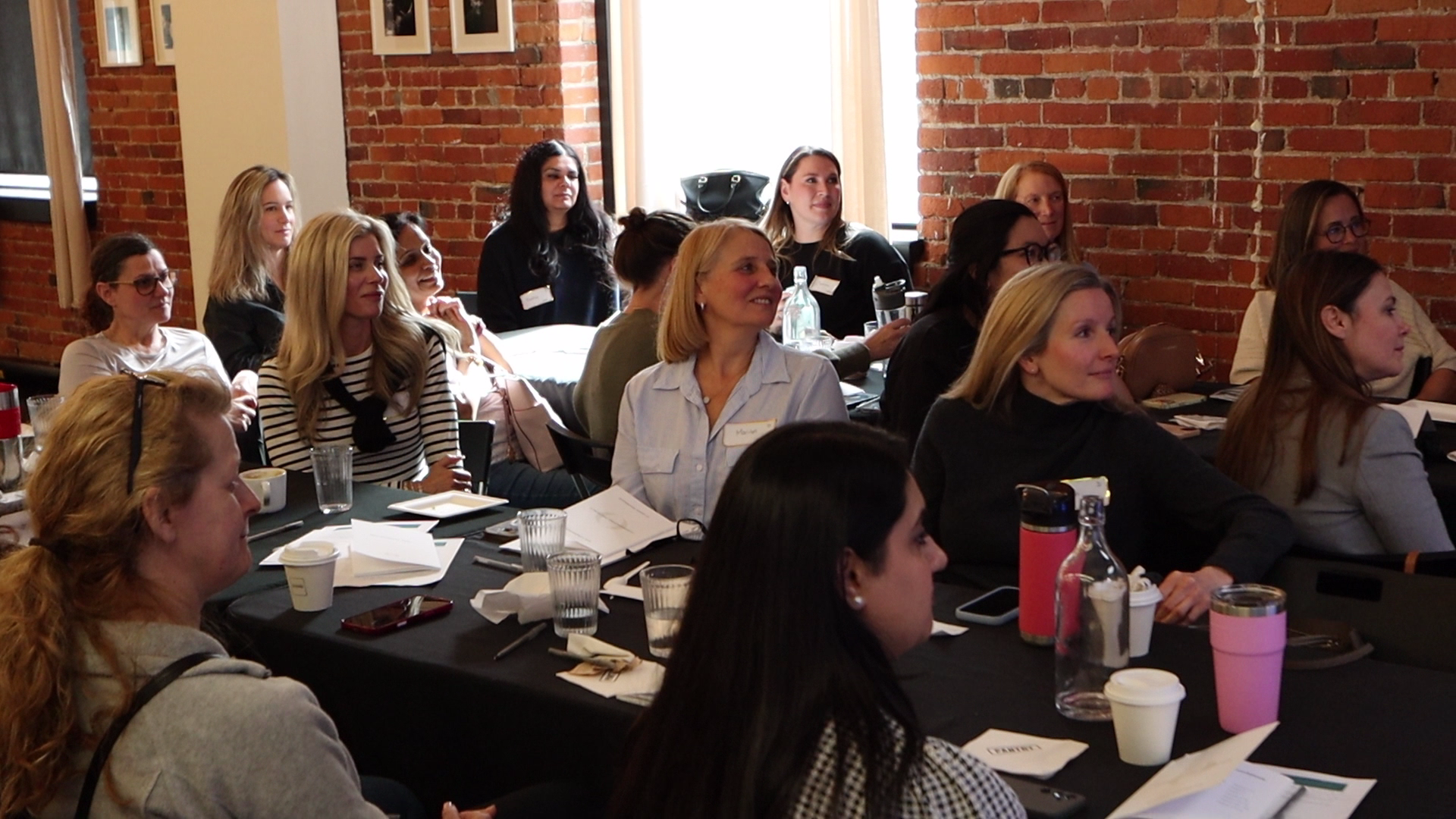 Group of women attending a seminar or workshop in a brick-walled room, sitting at tables with notebooks, drinks, and conference materials.