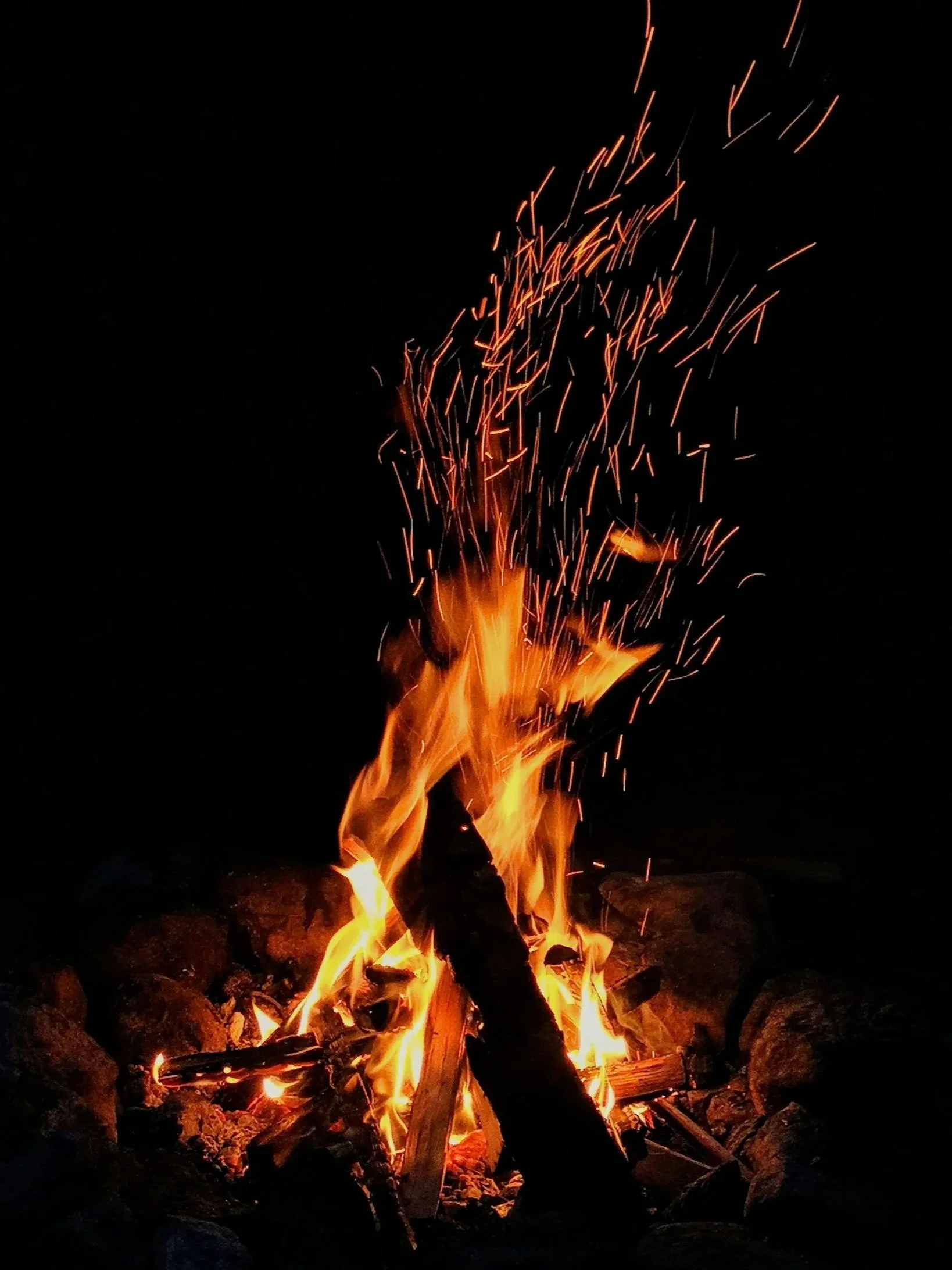 A campfire with orange flames and sparks rising into the night sky, surrounded by rocks.