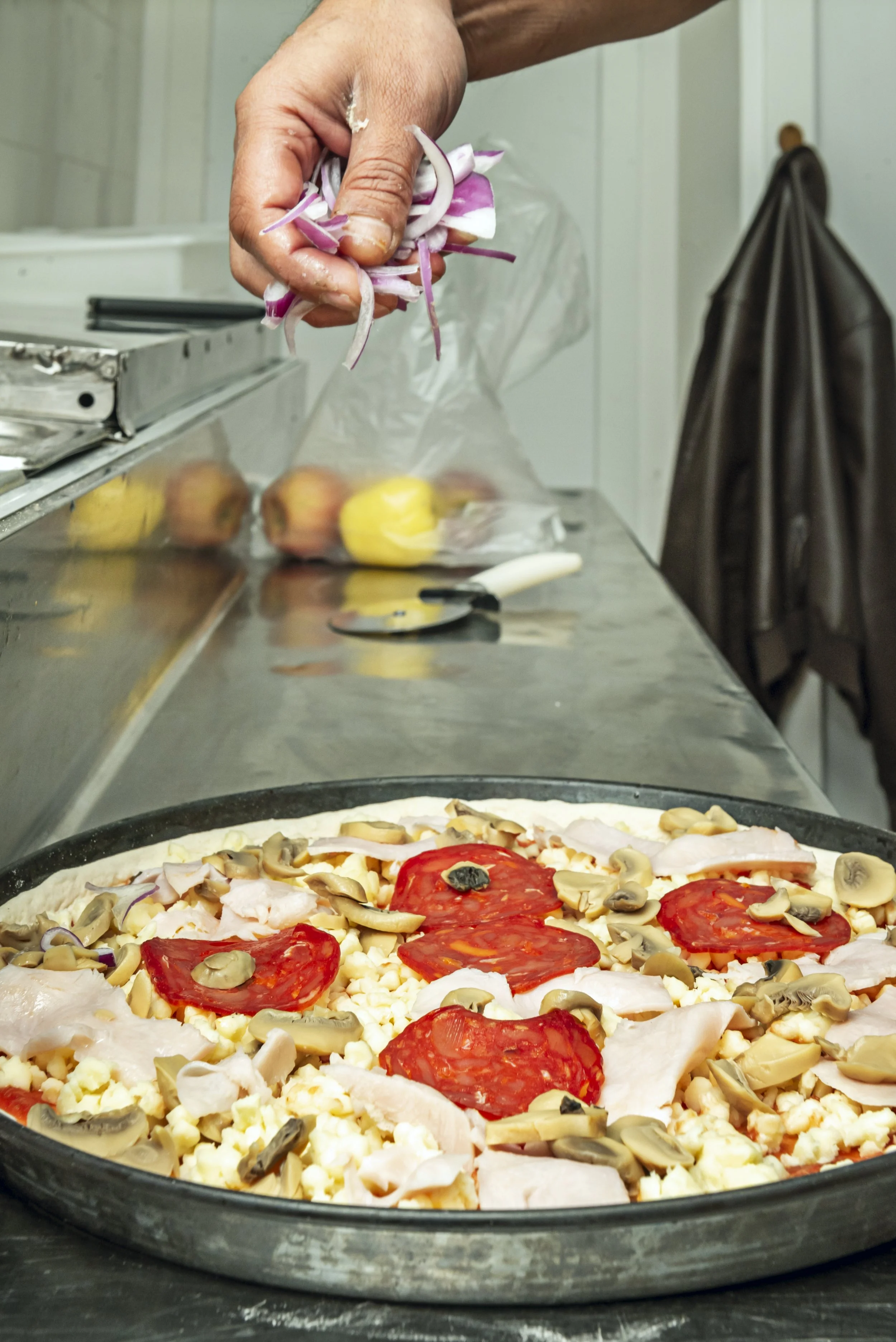 Persona añadiendo cebolla en una pizza antes de hornear, con ingredientes como tomate, pollo, queso y champiñones en una bandeja de pizza.