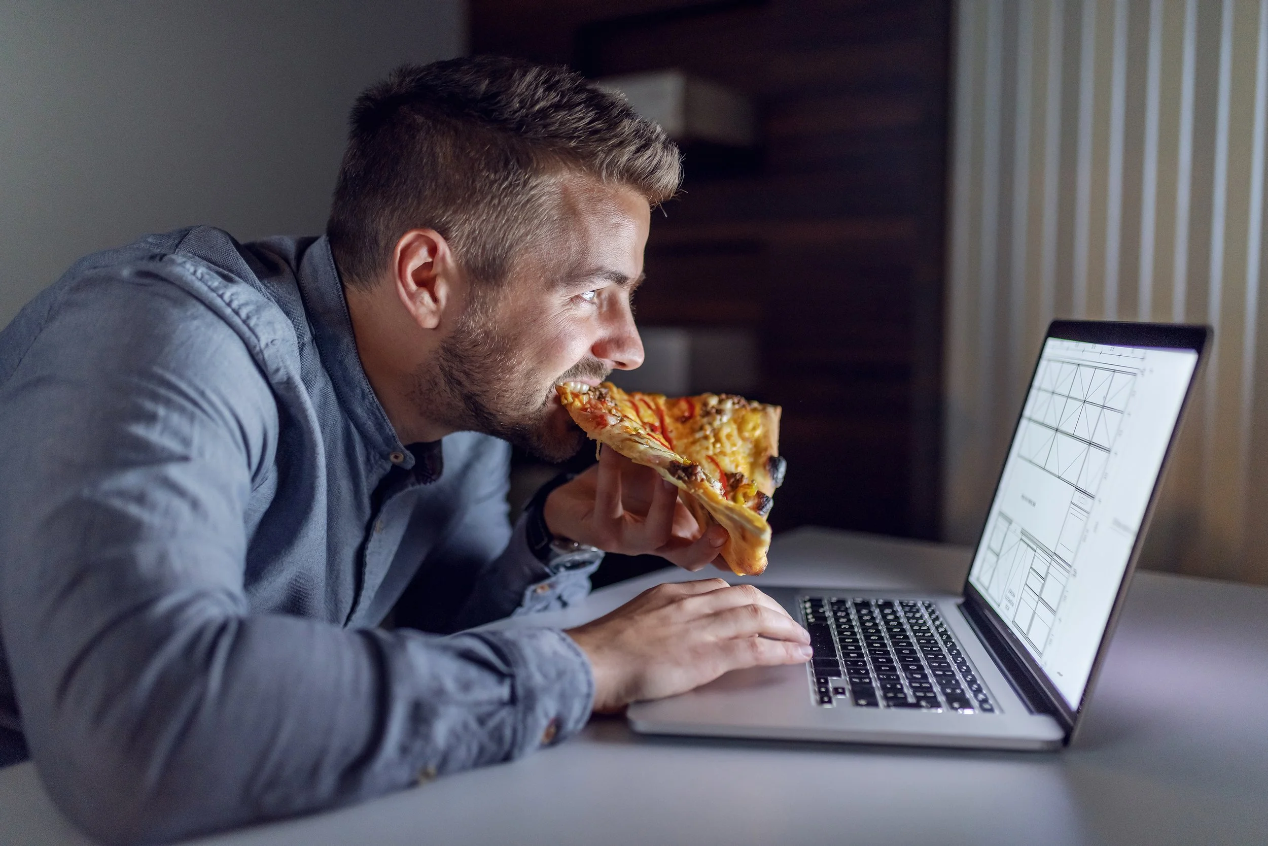 Joven comiendo pizza frente a una laptop en un entorno interior.