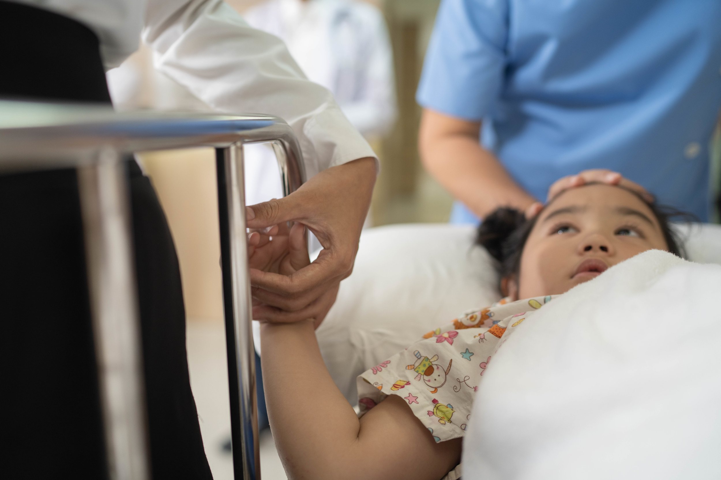 A young girl lying in a hospital bed, being attended to by medical staff. One staff member is holding her hand, and another appears to be checking her medical equipment.