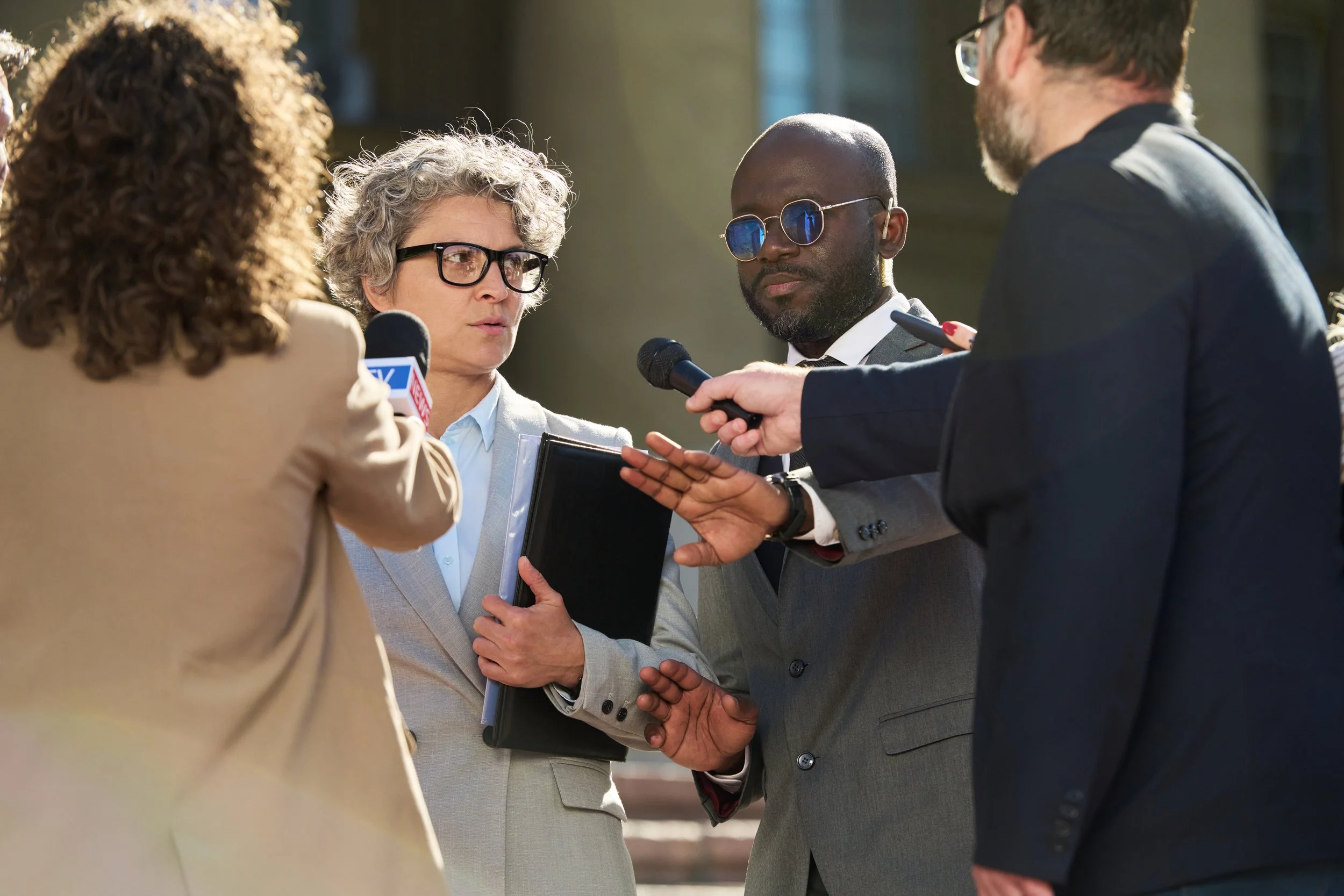 A woman with curly hair and glasses, holding a tablet, appears to be interviewing or speaking with two men, one with glasses and a dark suit, and another with a beard and glasses, outside a building.