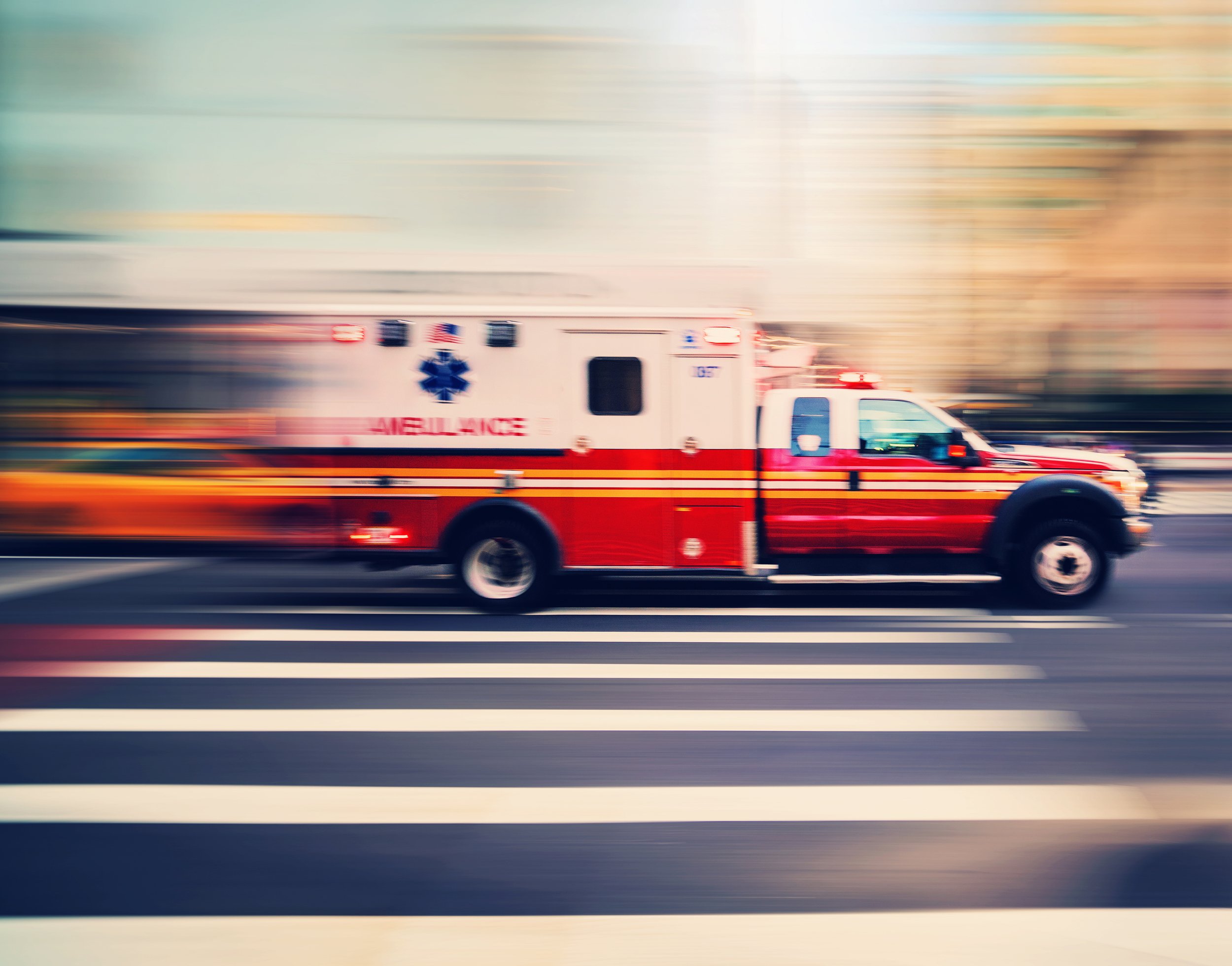 A moving ambulance driving on a road with painted lane lines, with a blurred cityscape in the background.