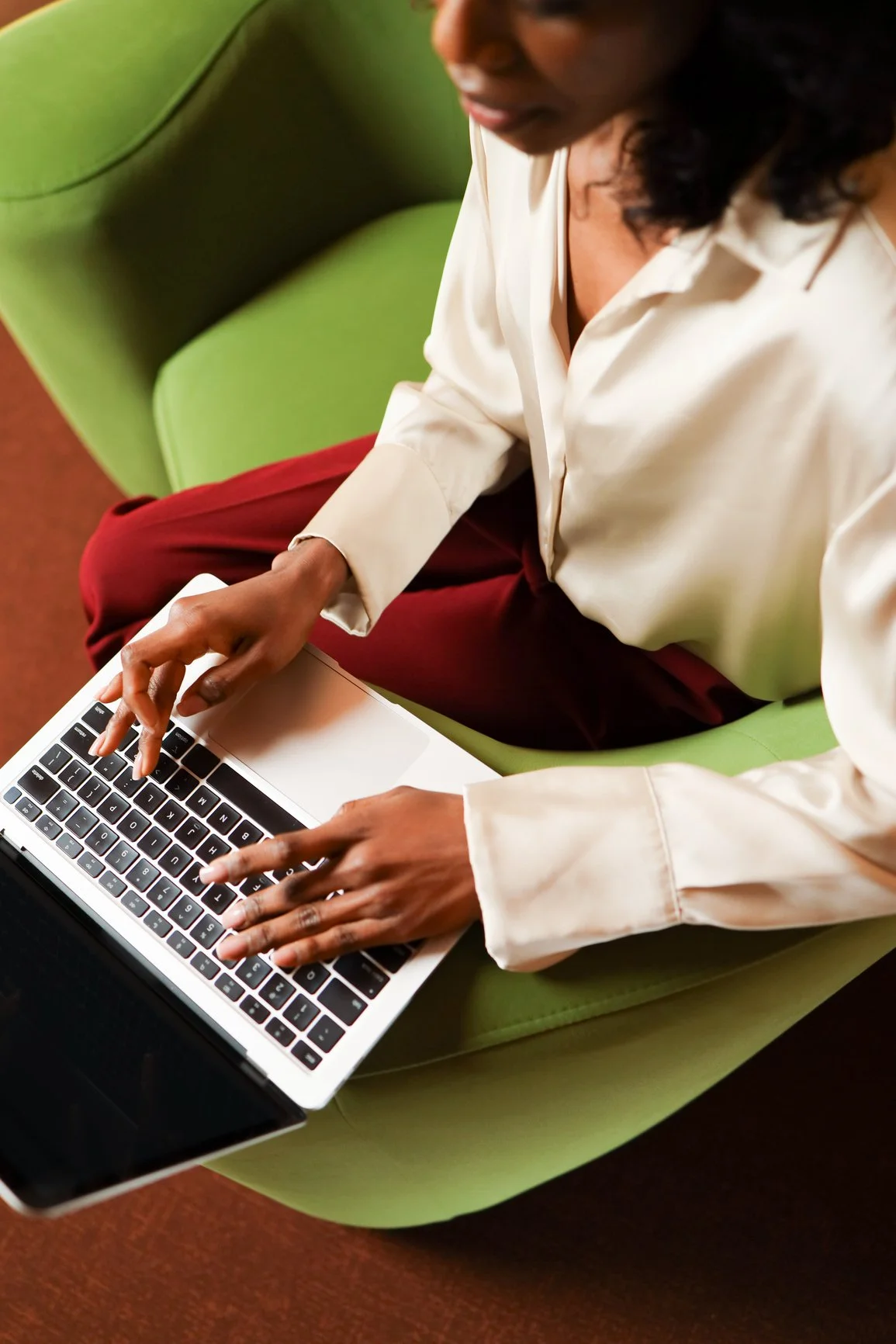 Overview of a woman in a cream blouse and burgundy pants typing on a laptop while seated in a green armchair.