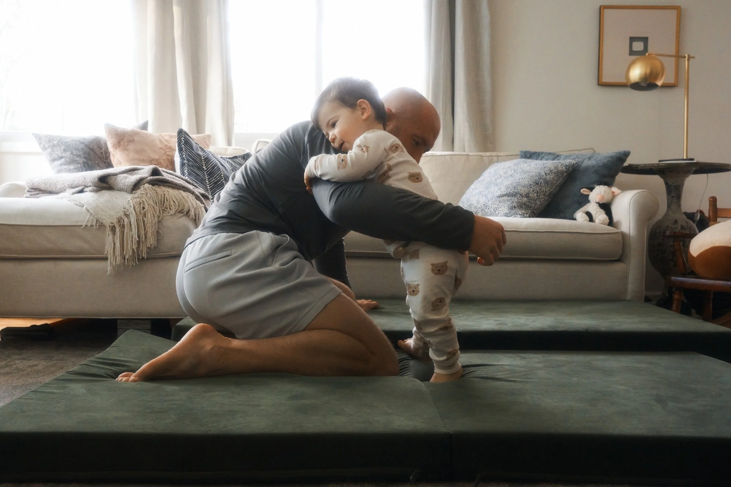 A man kneeling on a green mat, holding a young child in pajamas, helping them stand in a living room with a beige couch, pillows, and a stuffed animal in the background.