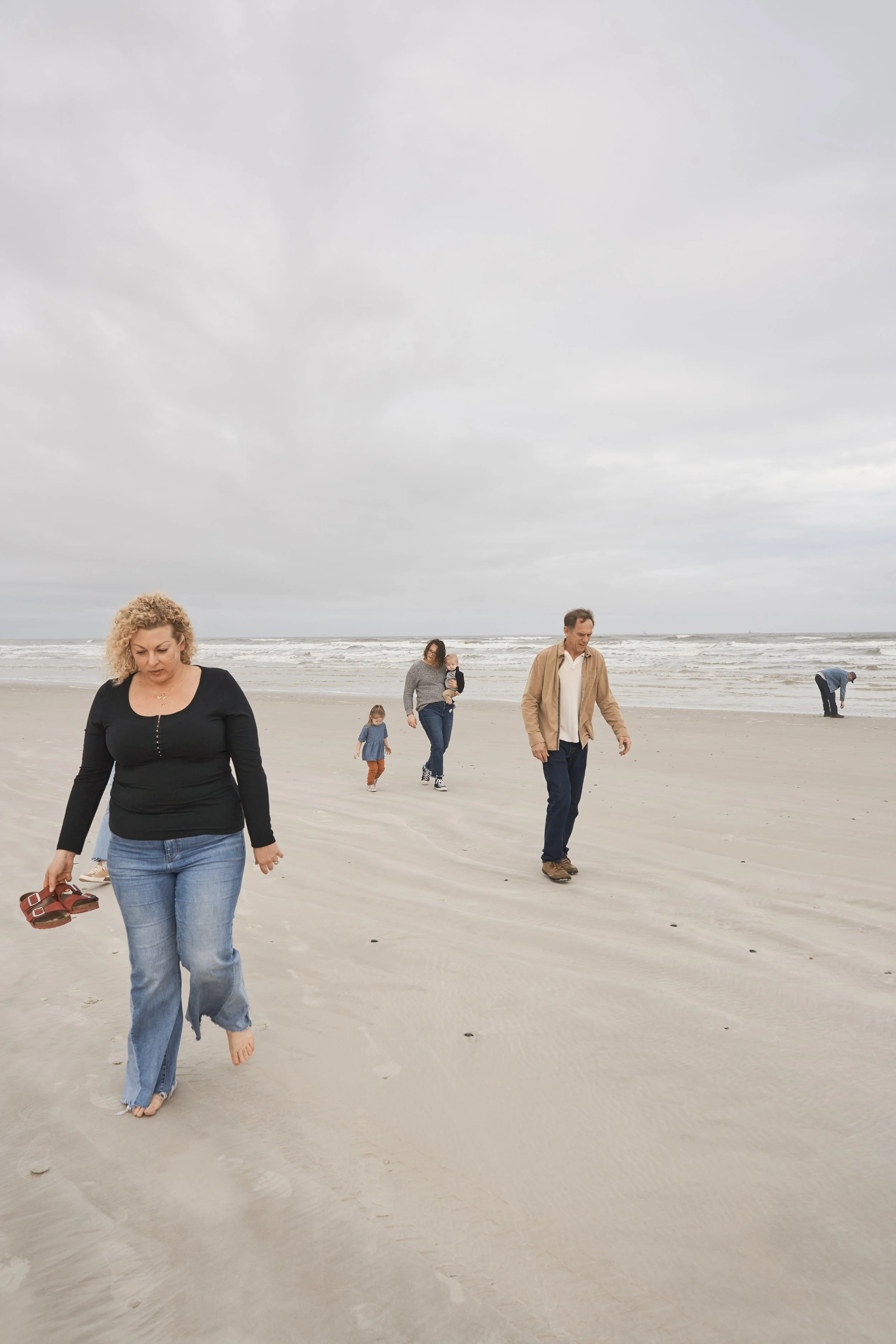 Family walking barefoot on the sandy beach under a cloudy sky with ocean waves in the background.