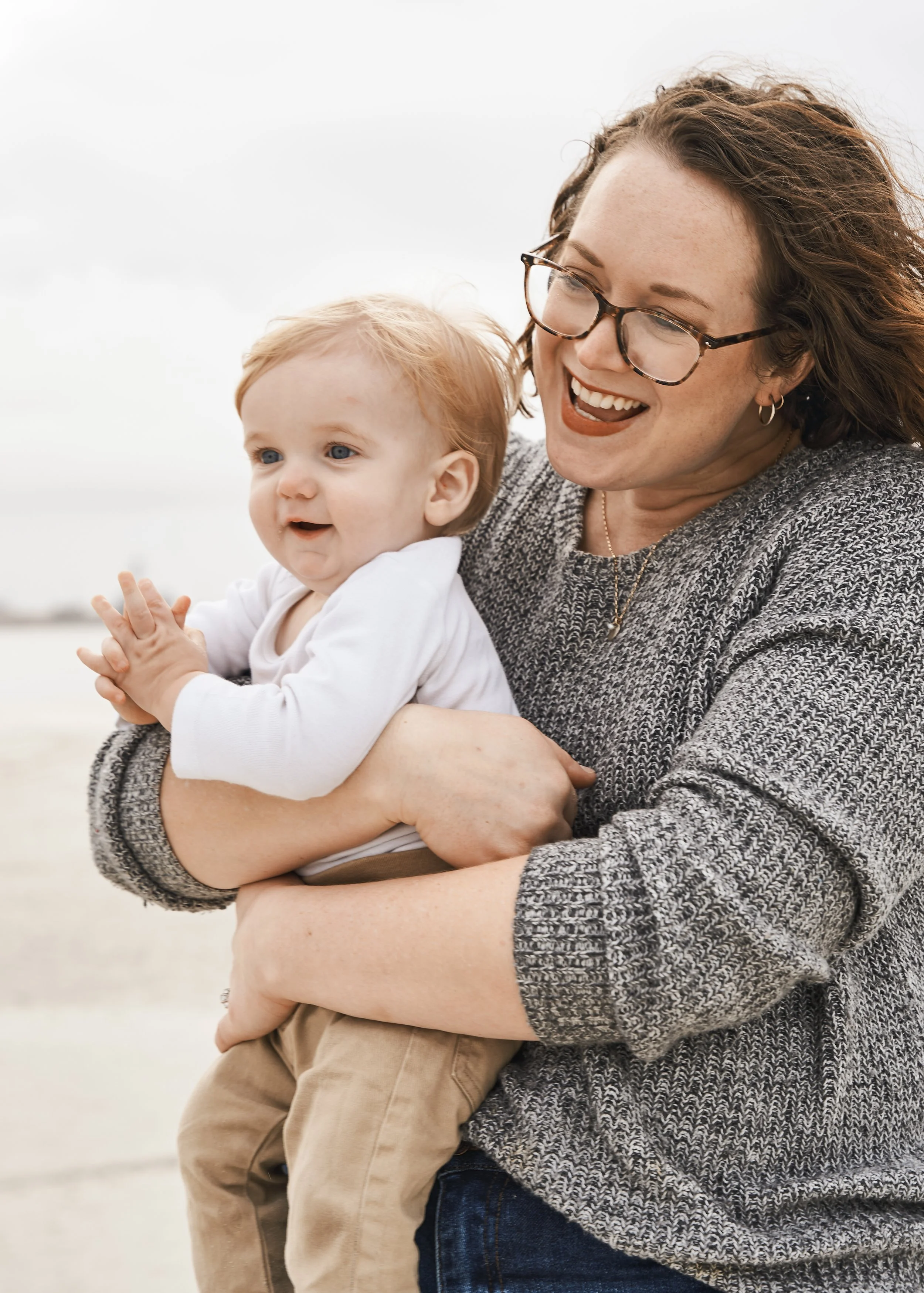 A woman with glasses holding a smiling toddler with blonde hair and blue eyes outdoors on a cloudy day.