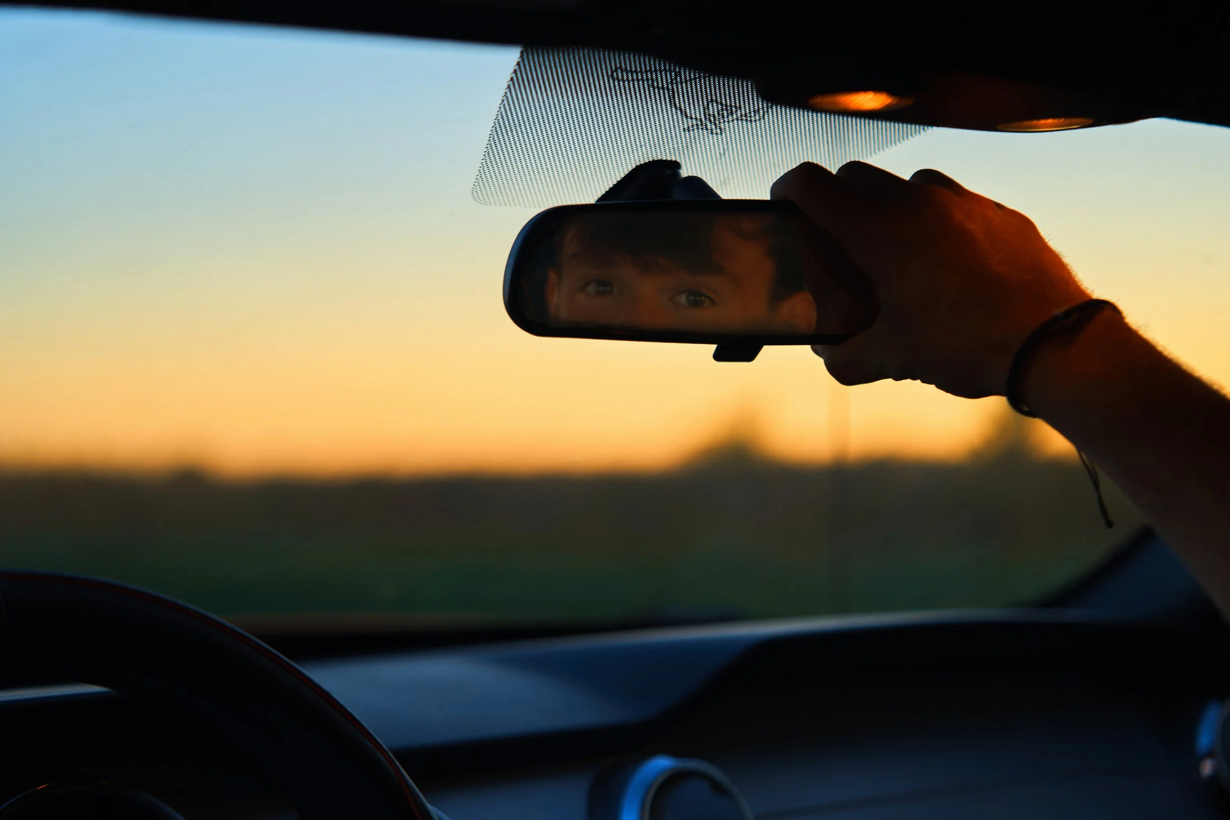 A person adjusting the rearview mirror in a car during sunset, with the reflection revealing their face and eyes.