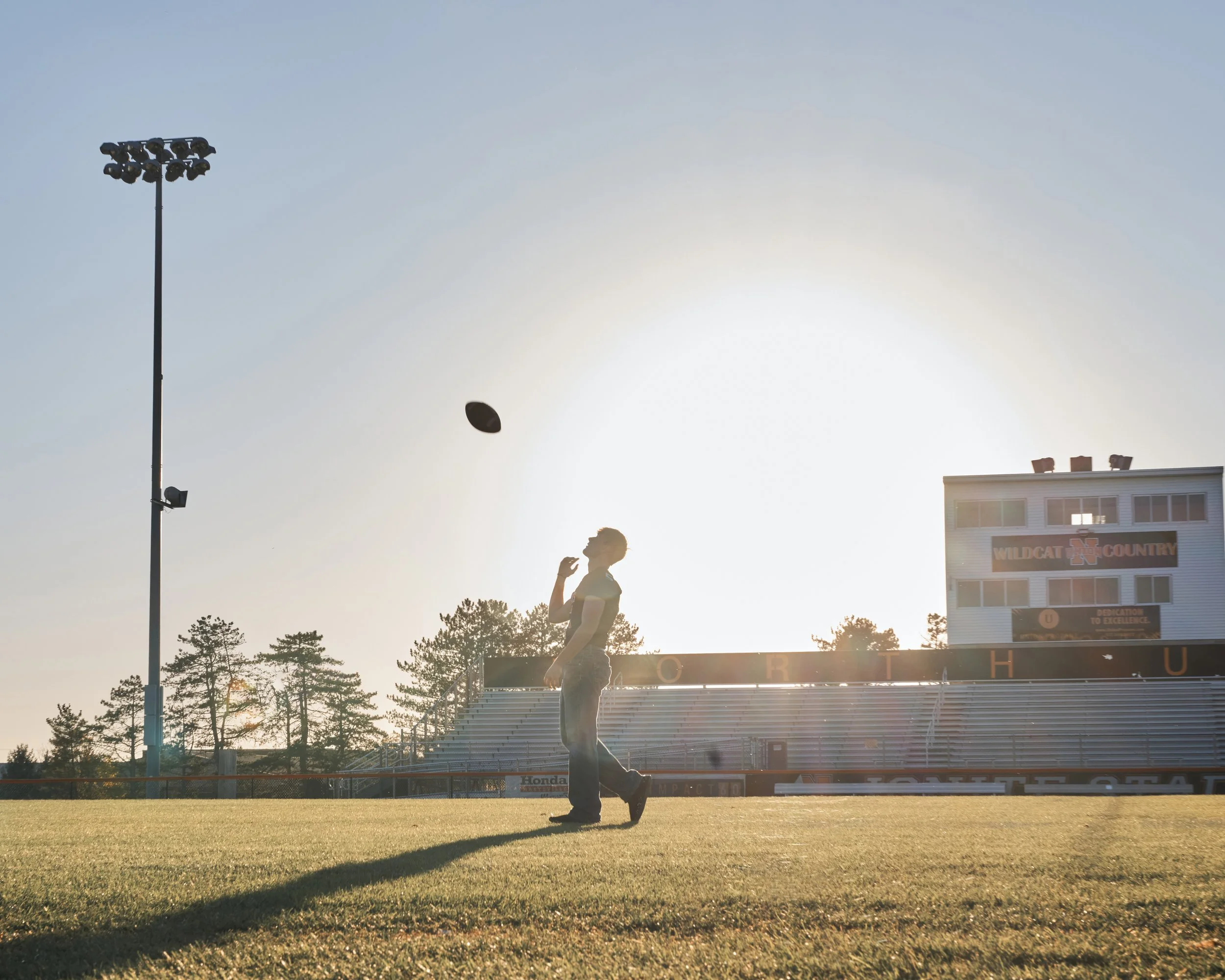 A person is football throwing on a field during sunset with trees and a stadium structure in the background.