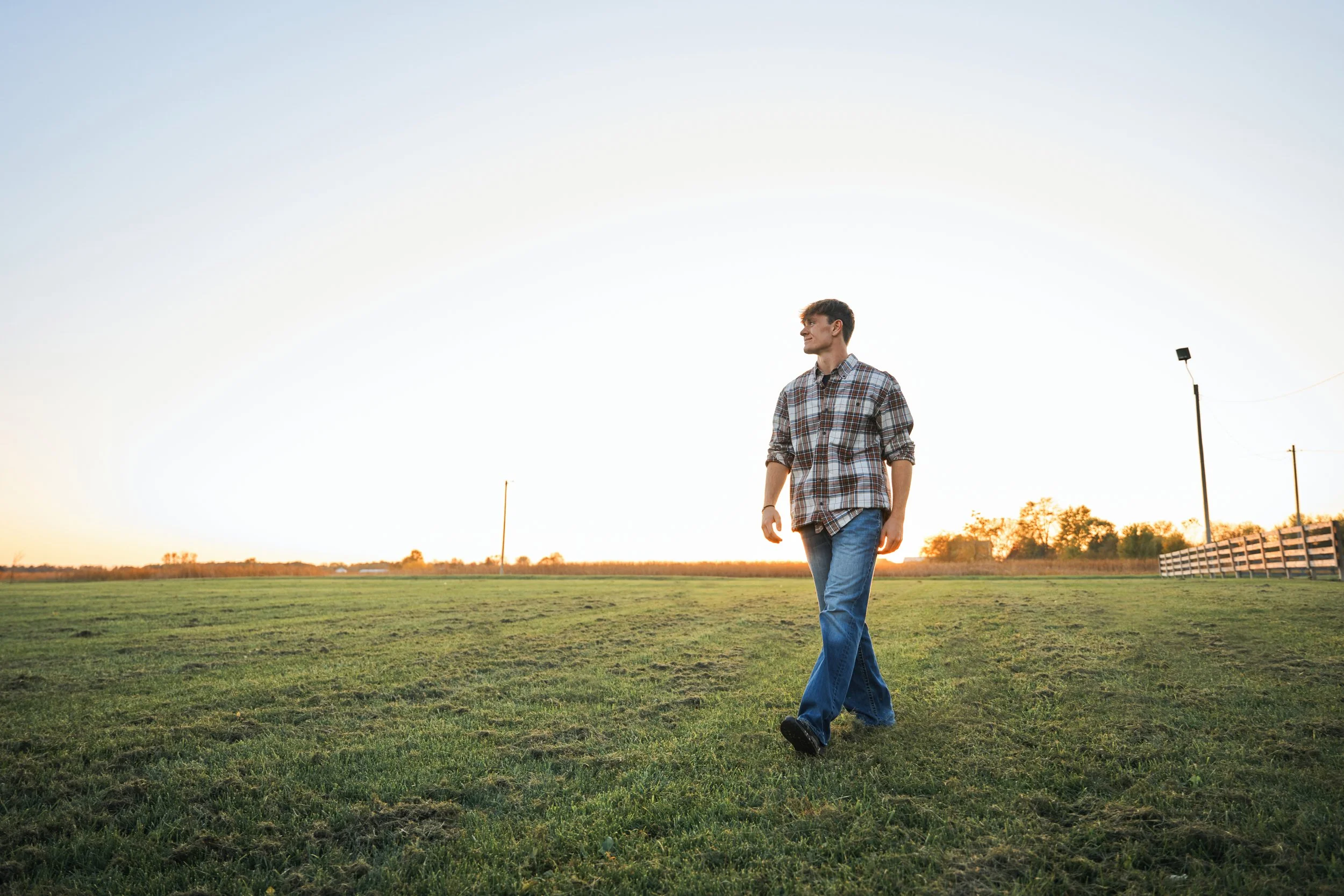 A young man in a plaid shirt and jeans walking across a grassy field during sunset.