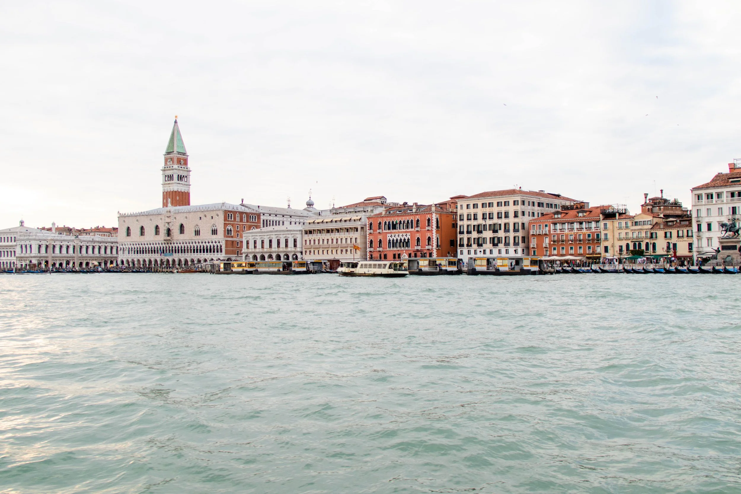 View of Venice, Italy, showing historic colorful buildings along the waterfront with boats docked and the famous St. Mark's Campanile in the background under a cloudy sky.