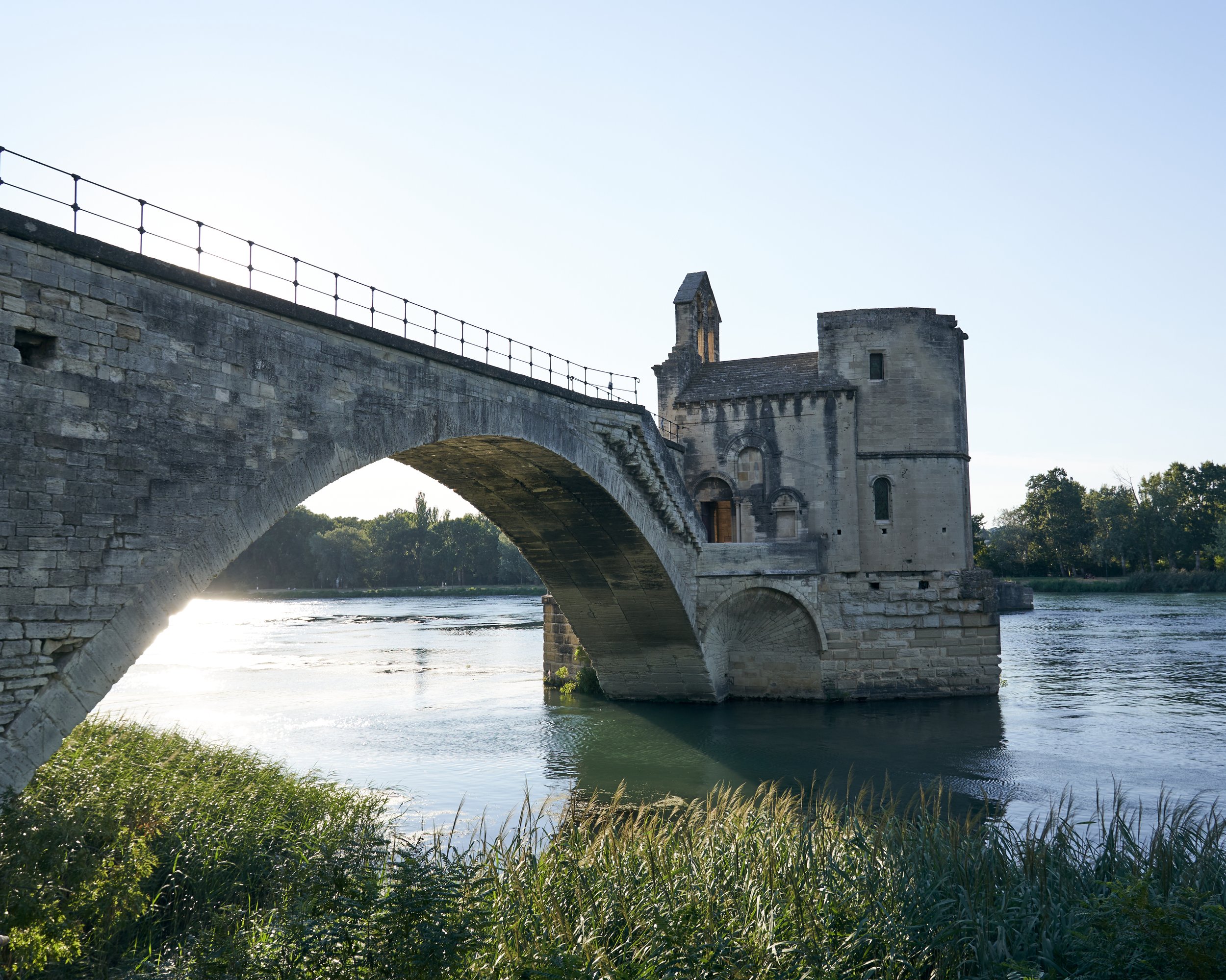 Historic stone bridge over a river with an ancient stone building attached, surrounded by green trees and grasses, under a clear blue sky.