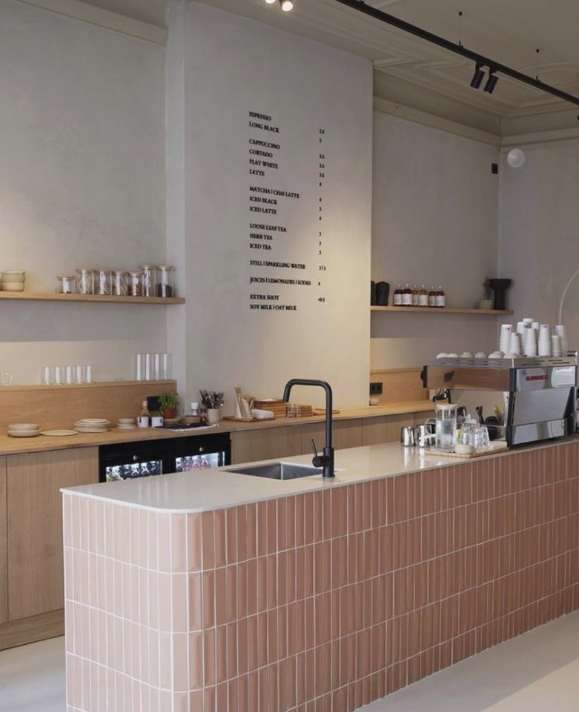A minimalist coffee shop counter with a white tiled front, light wooden shelves with coffee supplies, and a menu on the wall listing coffee drinks and prices.