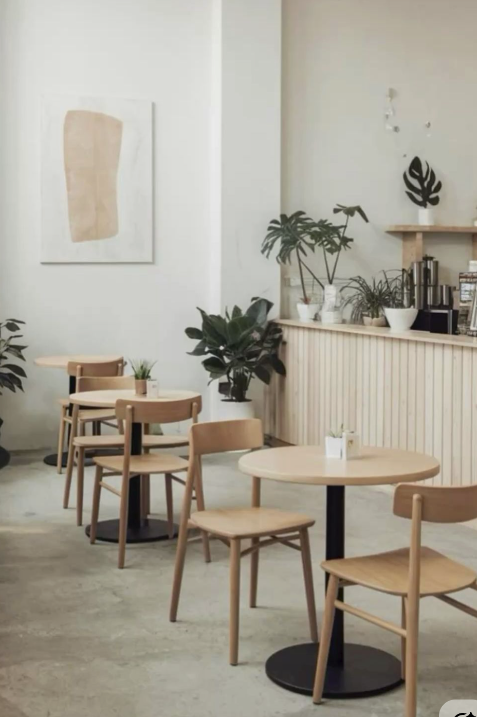 A cozy cafe interior with light wood tables and chairs, potted plants on the tables and counter, a framed abstract art piece on the wall, and a beverage station with coffee equipment in the background.