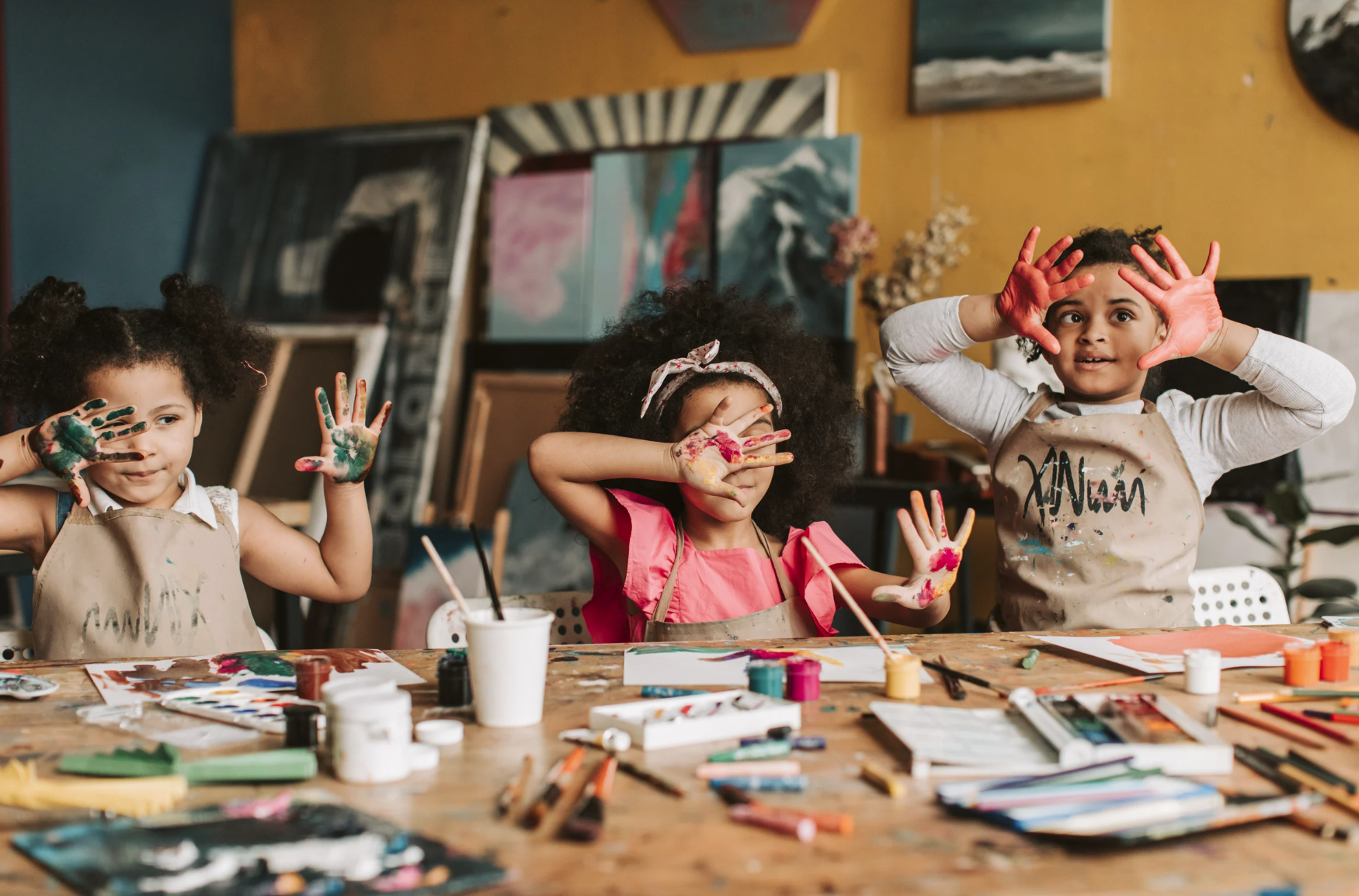Three young children with paint on their hands making playful gestures at a table covered with art supplies, in a colorful art studio.