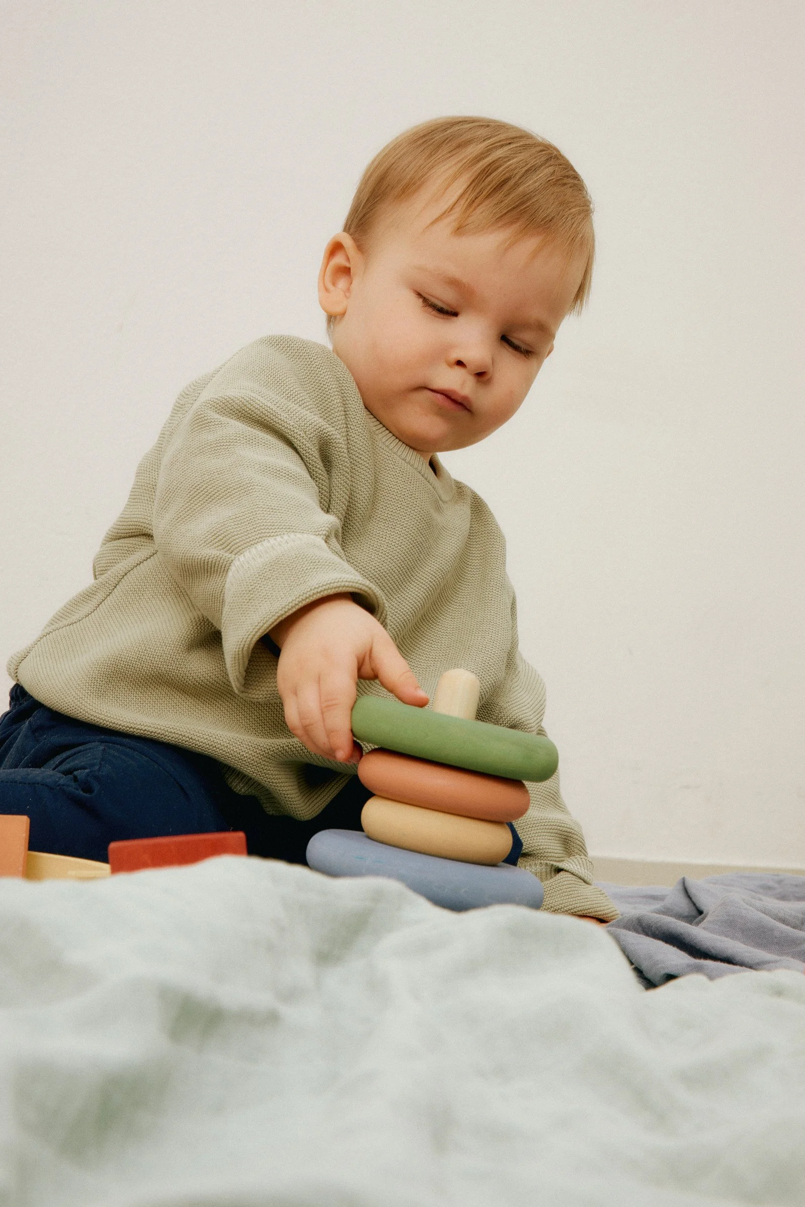 A young boy with blond hair wearing a beige long sleeve top and dark pants, sitting on a soft surface, stacking multicolored wooden rings in a tower.