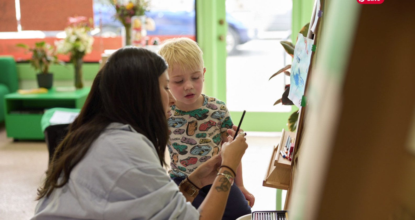 A woman with long dark hair, wearing a white shirt, showing a small paintbrush to a young boy with blonde hair wearing a colorful car-patterned shirt, inside a bright room with green and orange decor, artwork on a board, and a window with a door.