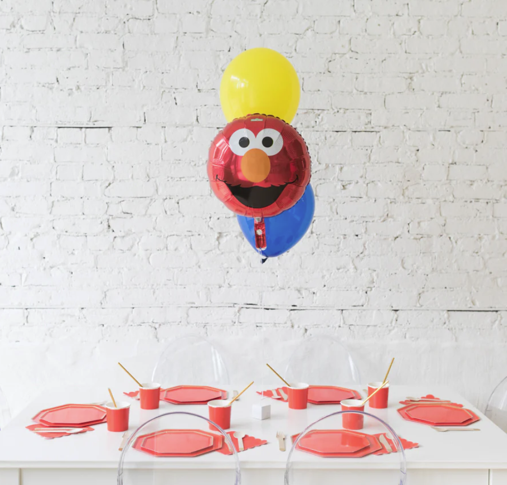 Table decorated for a Elmo-themed birthday party with red, yellow, blue balloons and Elmo character balloons, pink plates, cups with straws, and napkins.