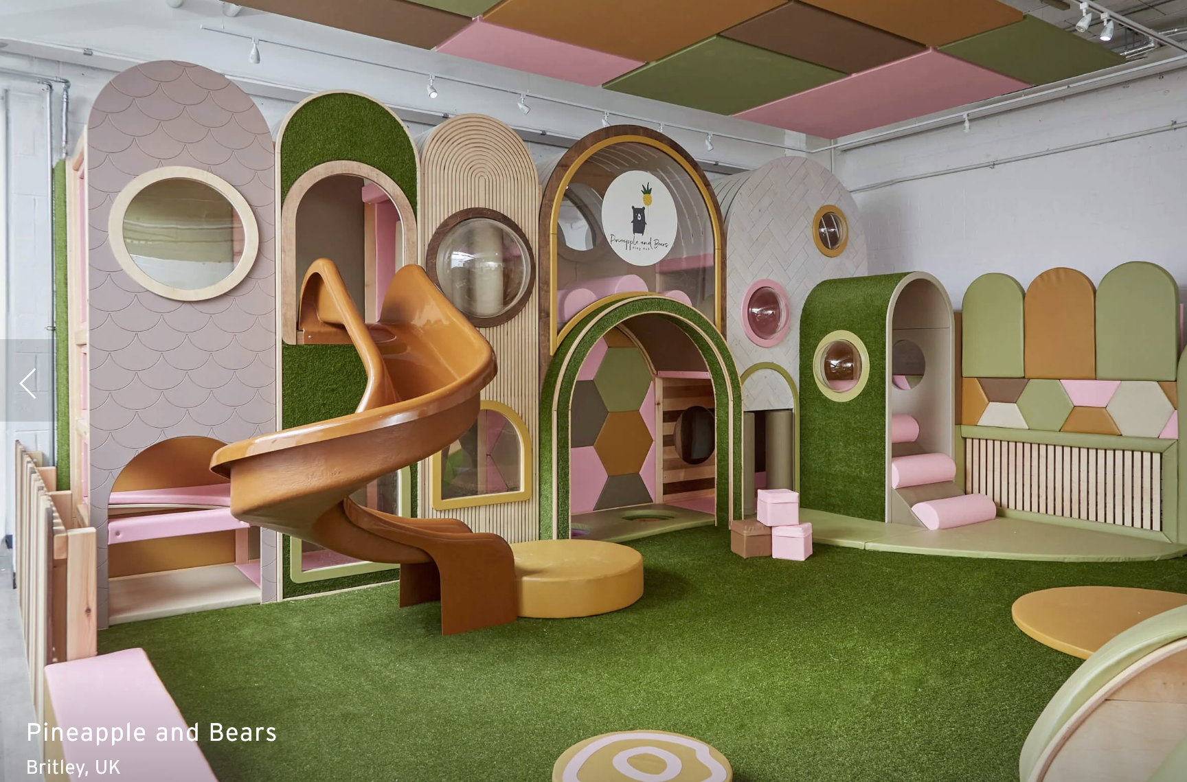 Colorful indoor play area with a green artificial turf floor, featuring various modular structures with arches, circular windows, a small slide, and cushioned steps in shades of pink, green, and orange.