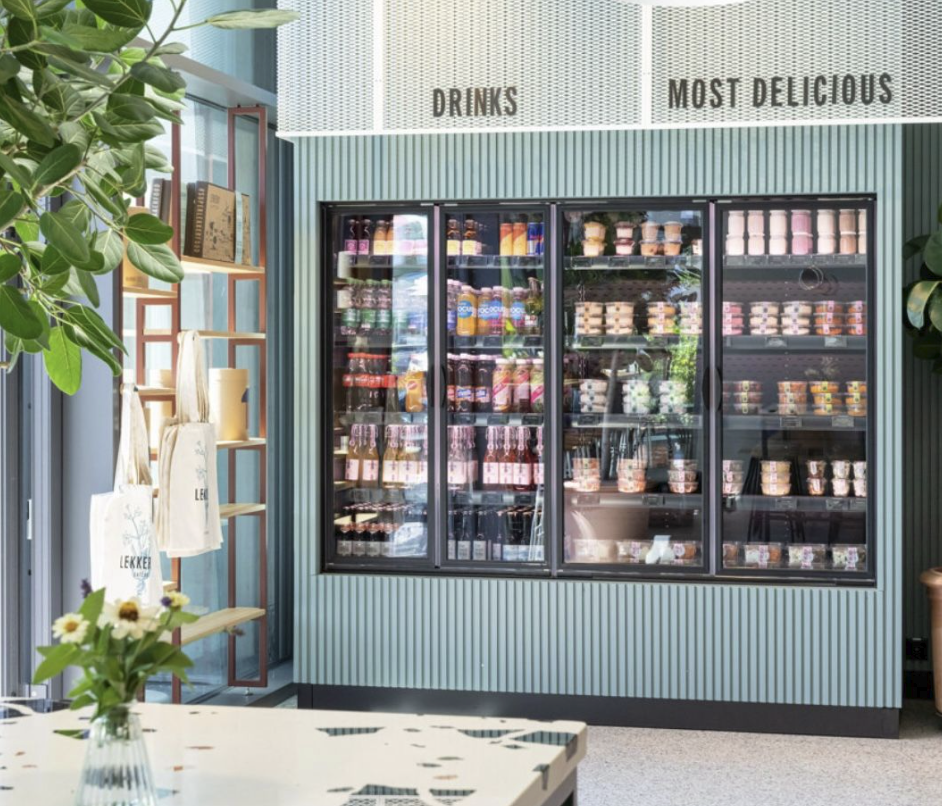 A store display with a glass refrigerator filled with various drinks and desserts, a shelf with tote bags, and a table with a vase of daisies in the foreground.